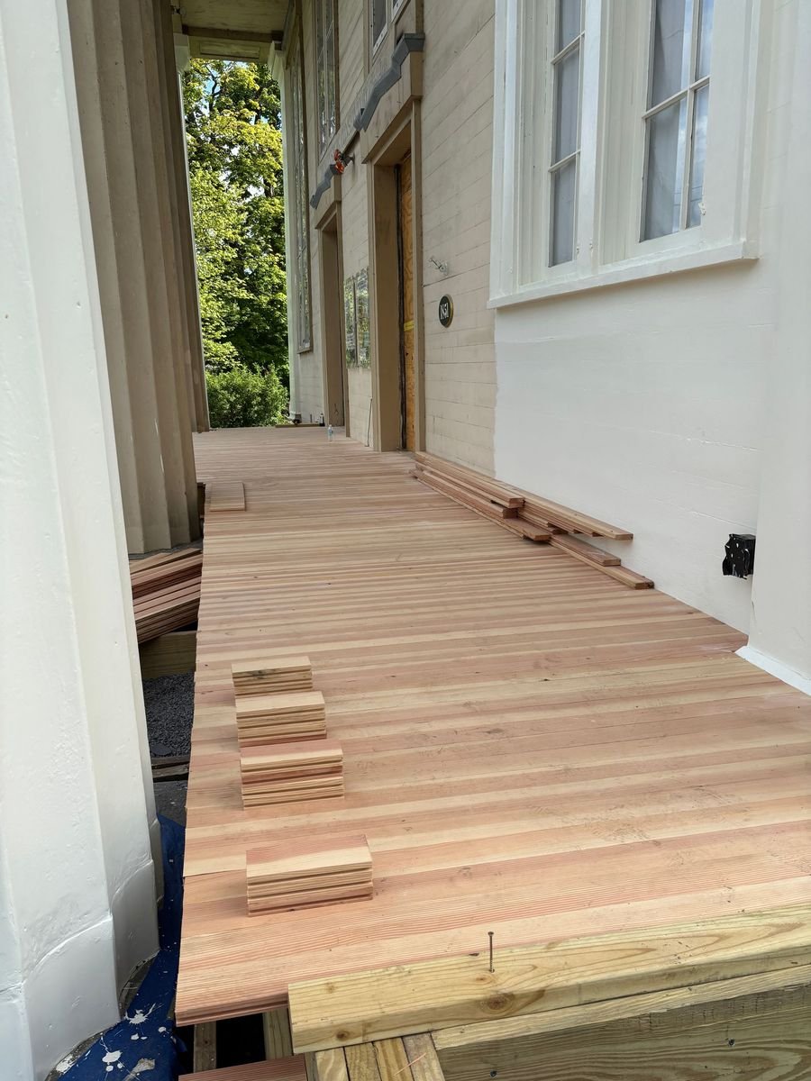 Wooden deck flooring construction with installed planks, some stacked nearby, on a porch attached to a house with white walls and large windows, surrounded by green trees.