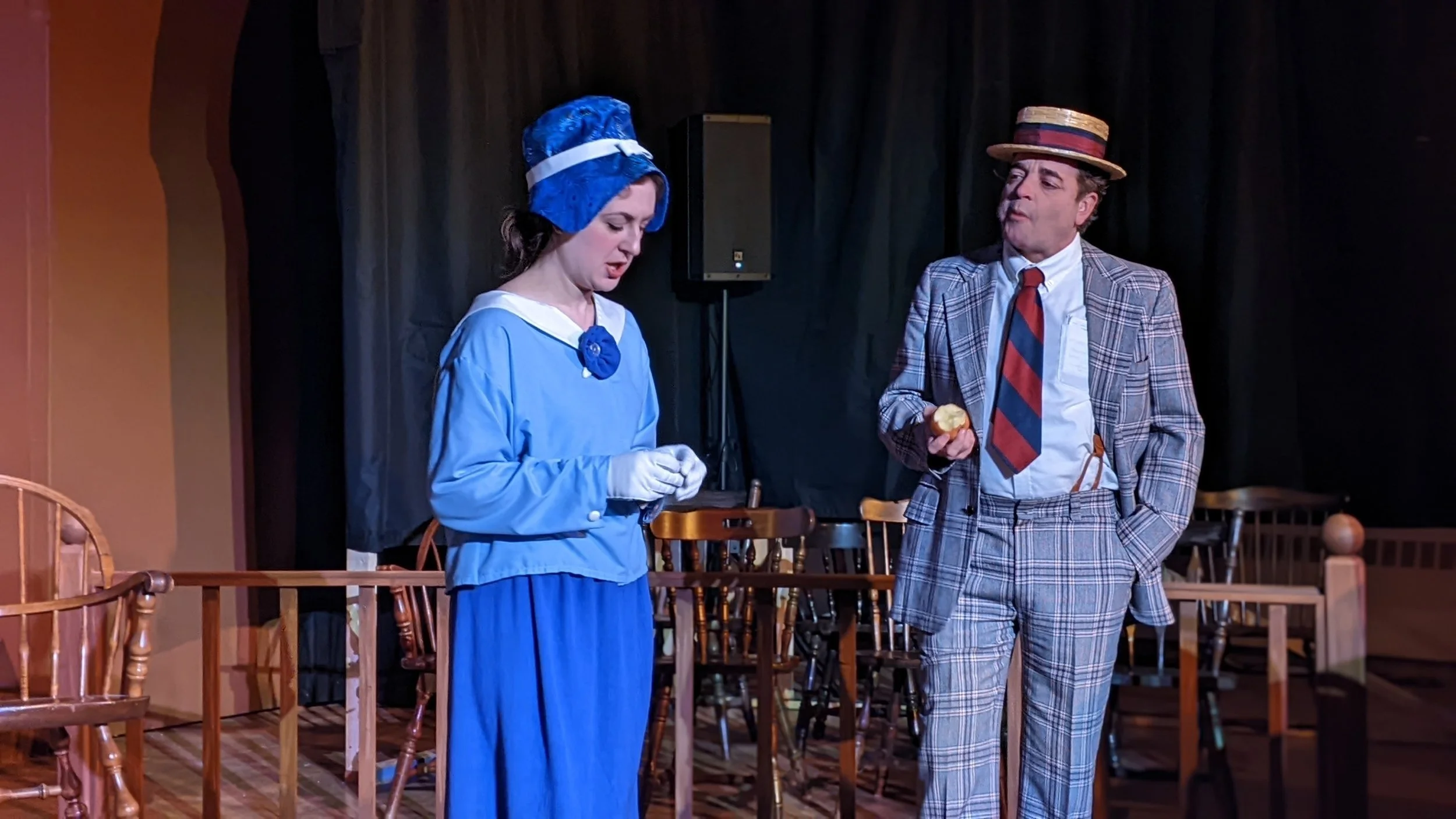 Two people dressed in vintage clothing stand on a stage with wooden chairs and a black curtain in the background, performing in a play.