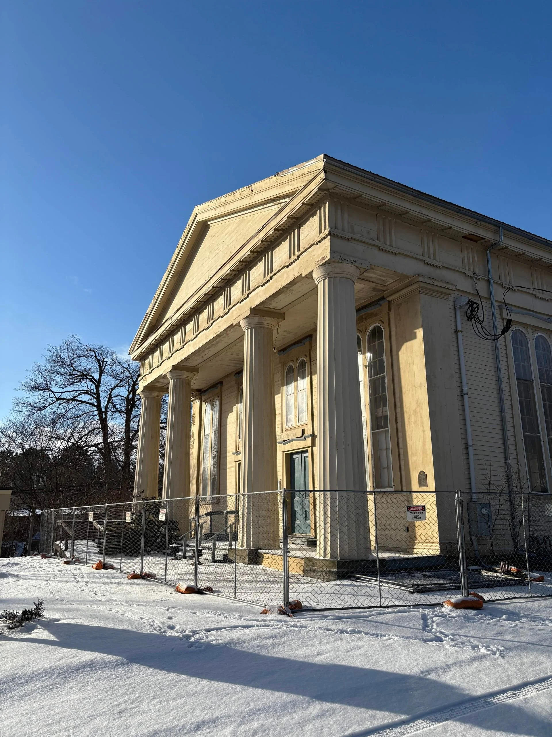 Exterior of thConservatory building with tall columns, surrounded by a construction fence. The building has a pediment, decorative moldings, and windows. Snow covers the ground, and the sky is clear blue. There are leafless trees in the background.