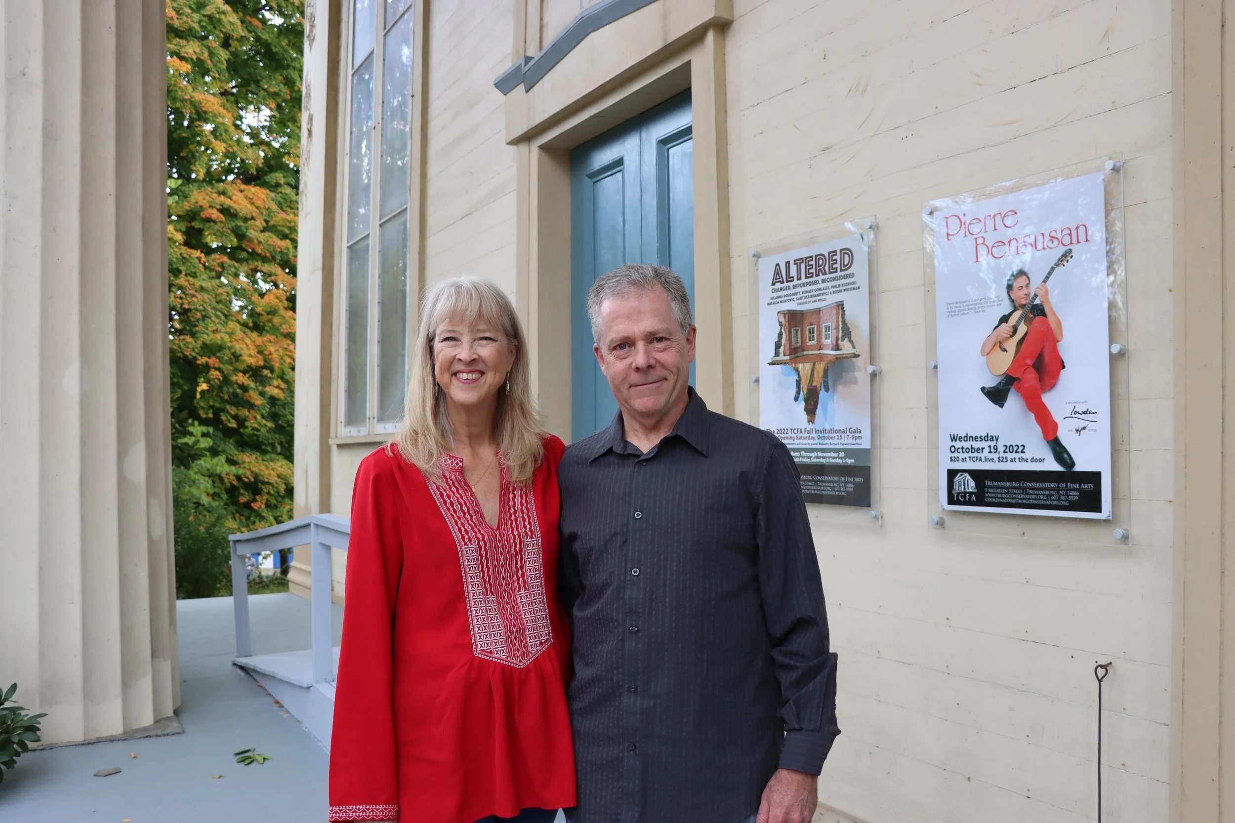 Two people, a woman with long blonde hair wearing a red embroidered blouse and a man with short gray hair wearing a black shirt, are standing outside in front of a beige building with posters on the wall. The background features a tree with green and orange leaves, indicating fall.