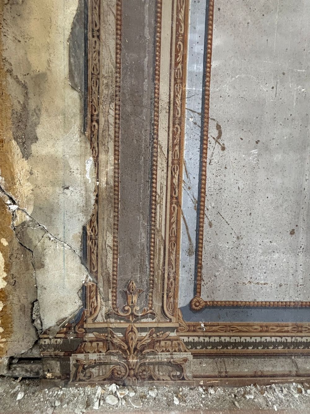 Fragment of a vintage decorative tile floor with ornate brown and beige borders, damaged with cracked and missing tiles, and exposed concrete underneath.