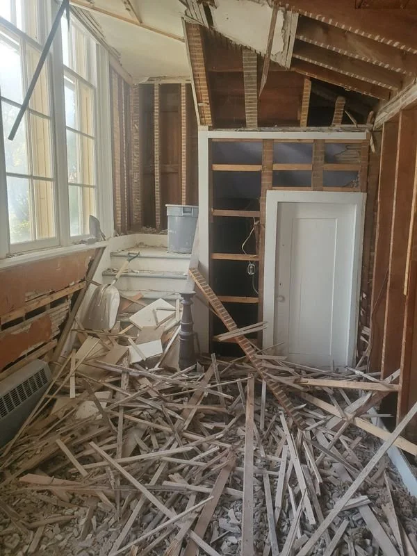 Room with damaged ceiling and collapsed wood, debris covering the floor, partially exposed walls, and a closed white door.