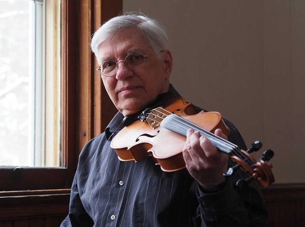 An elderly man with gray hair and glasses holding a violin near his shoulder in a warmly lit room with a window in the background.
