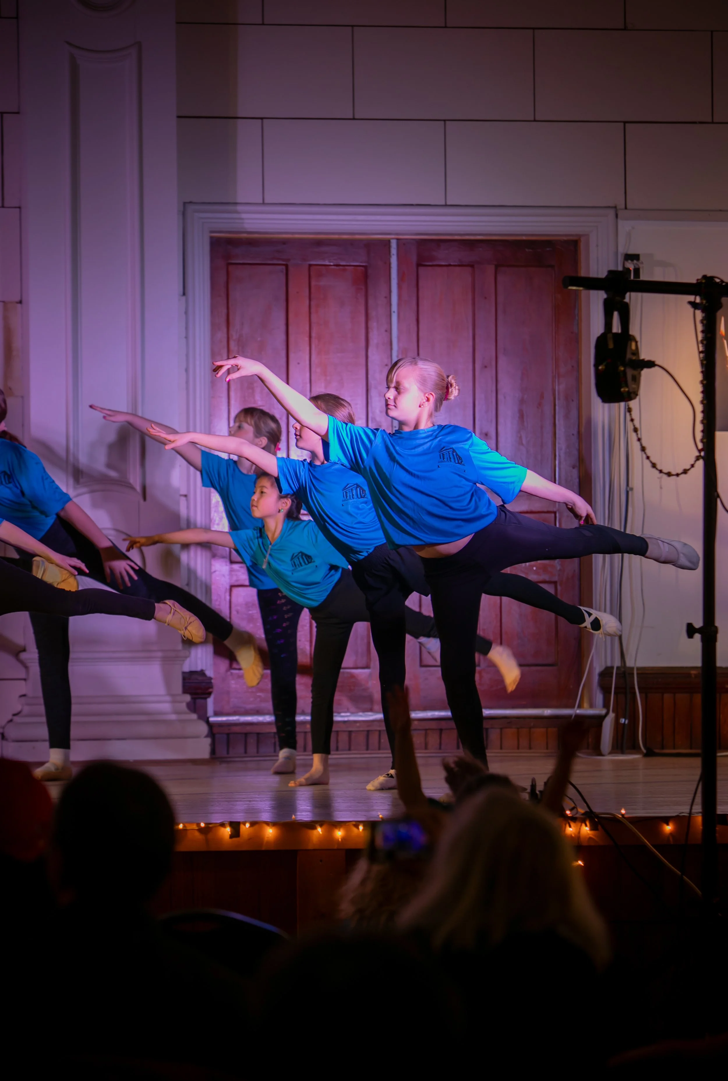 Children dressed in blue shirts performing ballet on stage, with audience watching.