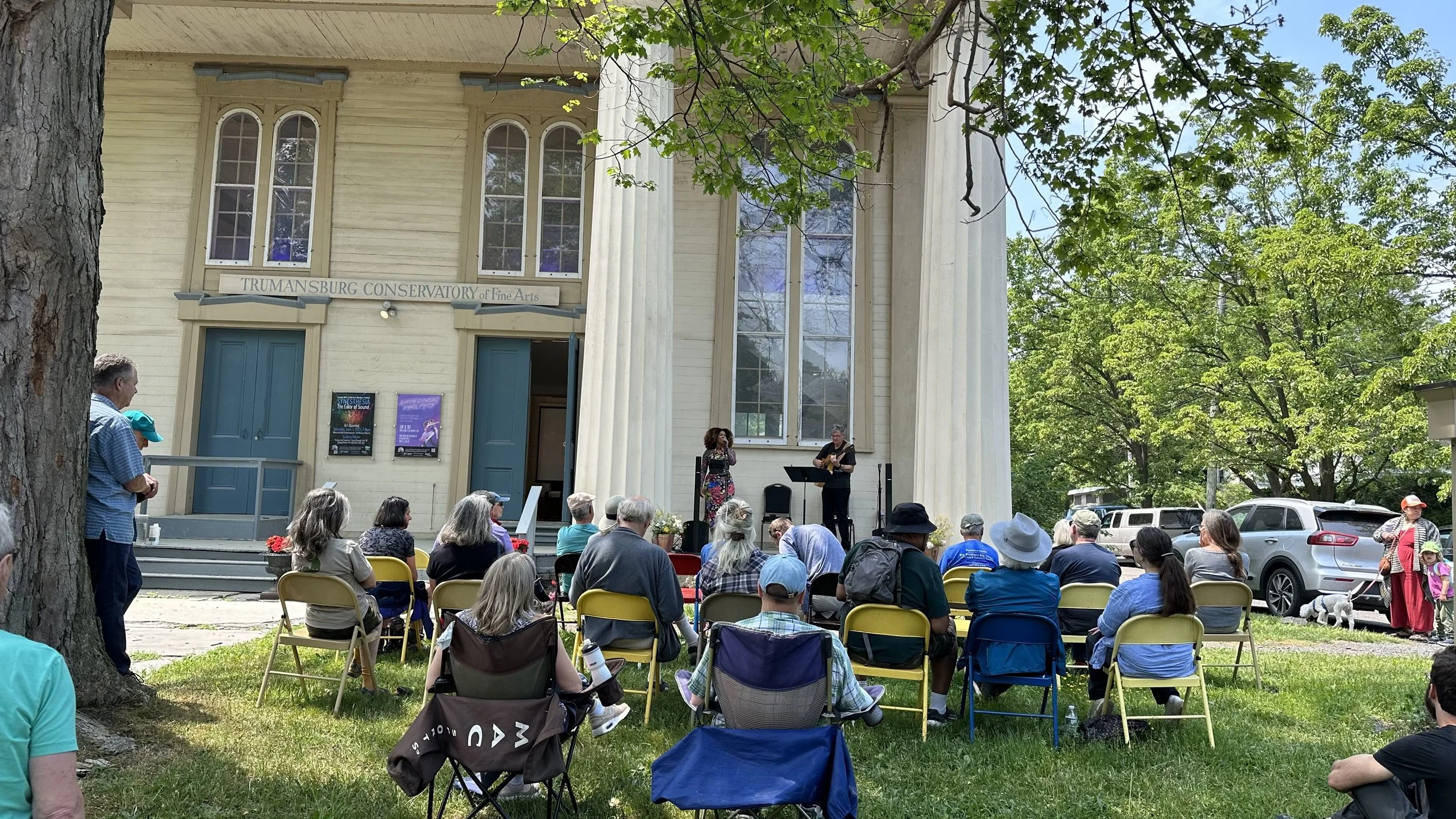 A crowd gathered outside the Trumansburg Conservatory of Fine Arts to watch a musical performance with a female singer and a male guitarist. The building has large windows and a sign above the door, with trees and parked cars in the background.