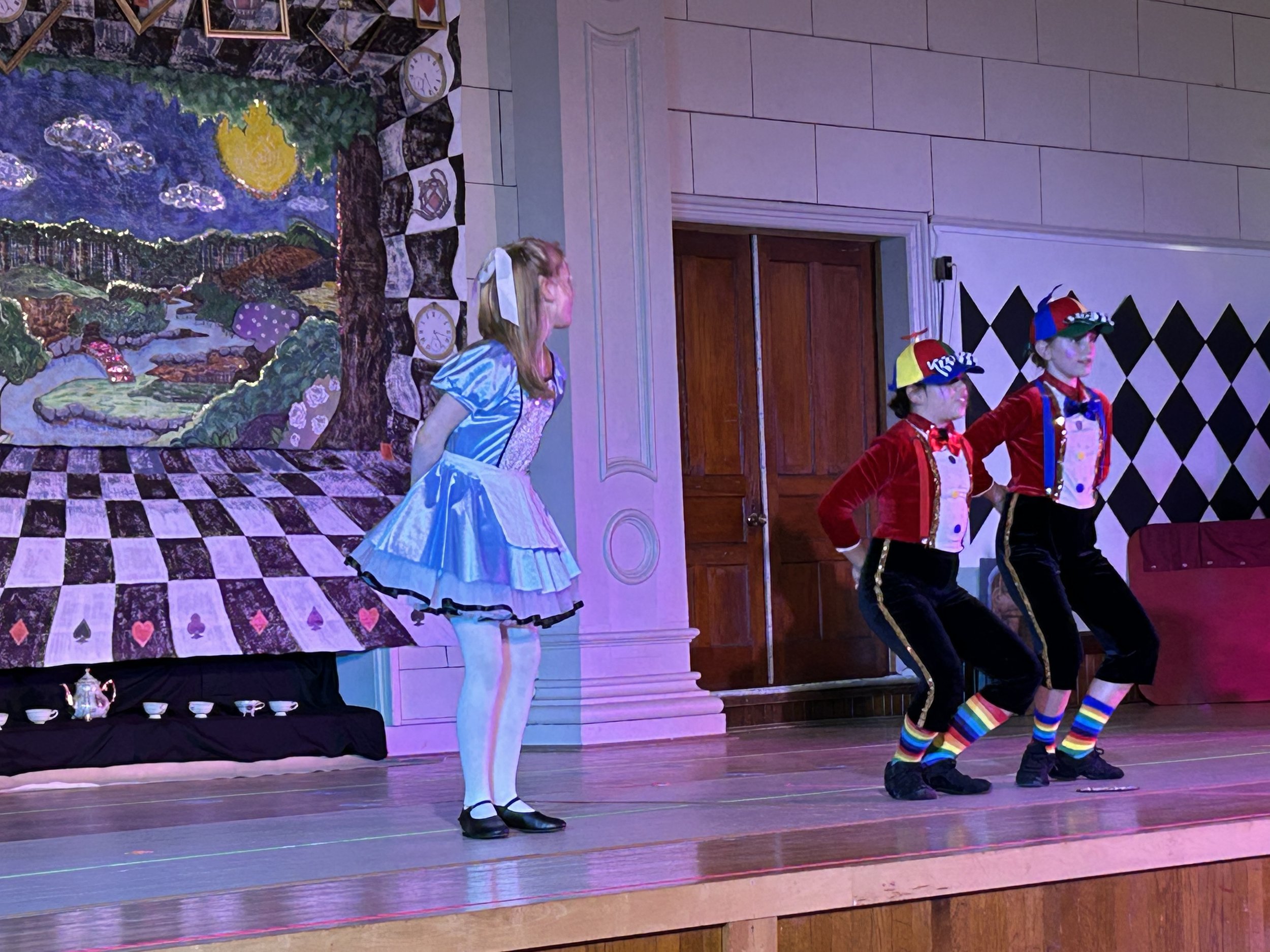 Children in colorful costumes performing on stage as part of a play, with a backdrop depicting a fantasy landscape with a large moon, clouds, and trees.