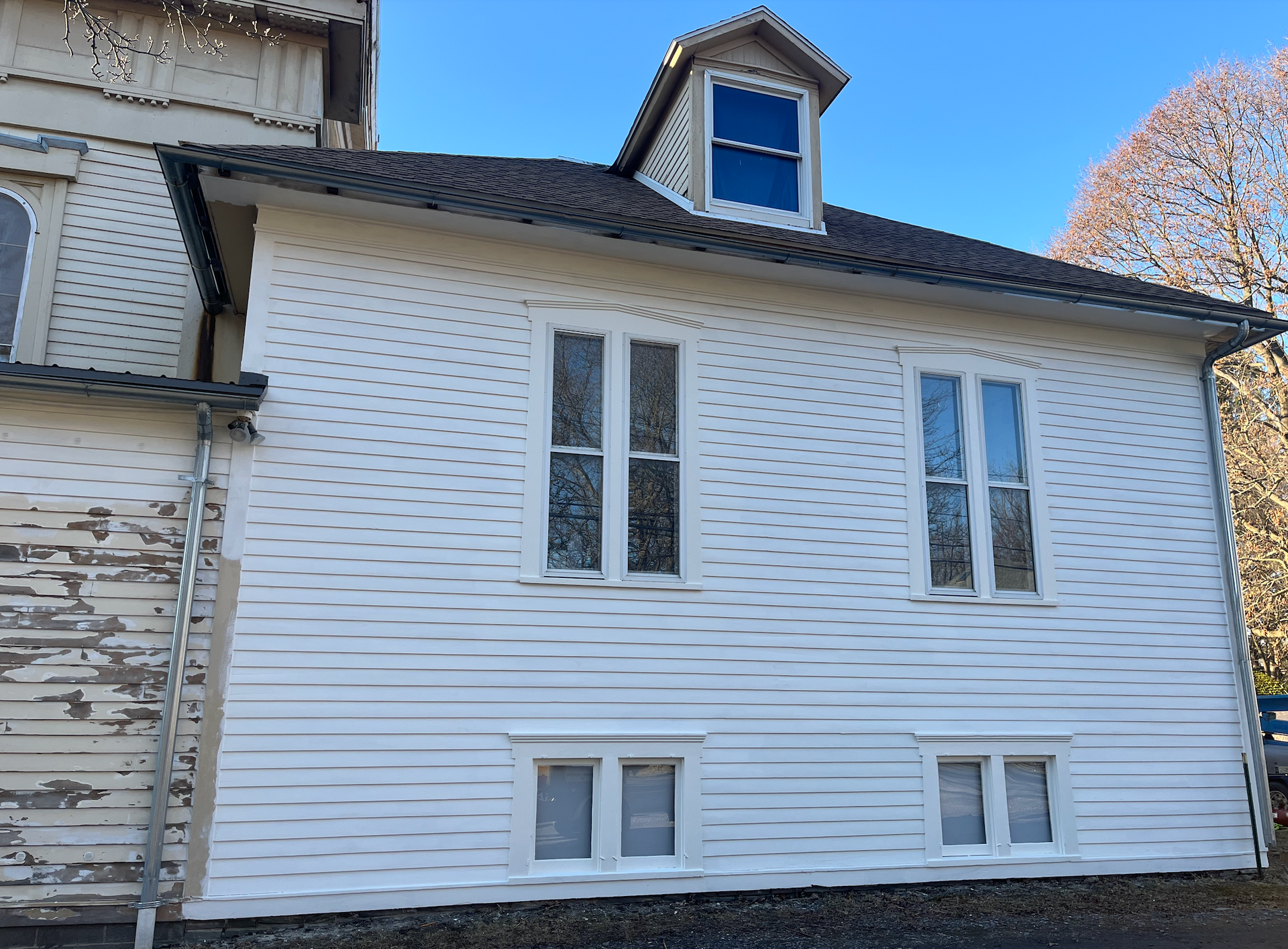 The image shows the side of TCFA with white vinyl siding. The building has a gable roof with three windows: two standard windows and a small dormer window. There is a black gutter along the roof edge and a downspout on the left side.