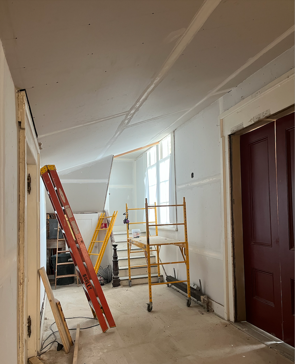 Interior of TCFA under renovation with scaffolding, a ladder, and construction materials, featuring a staircase and a window with sunlight coming in.