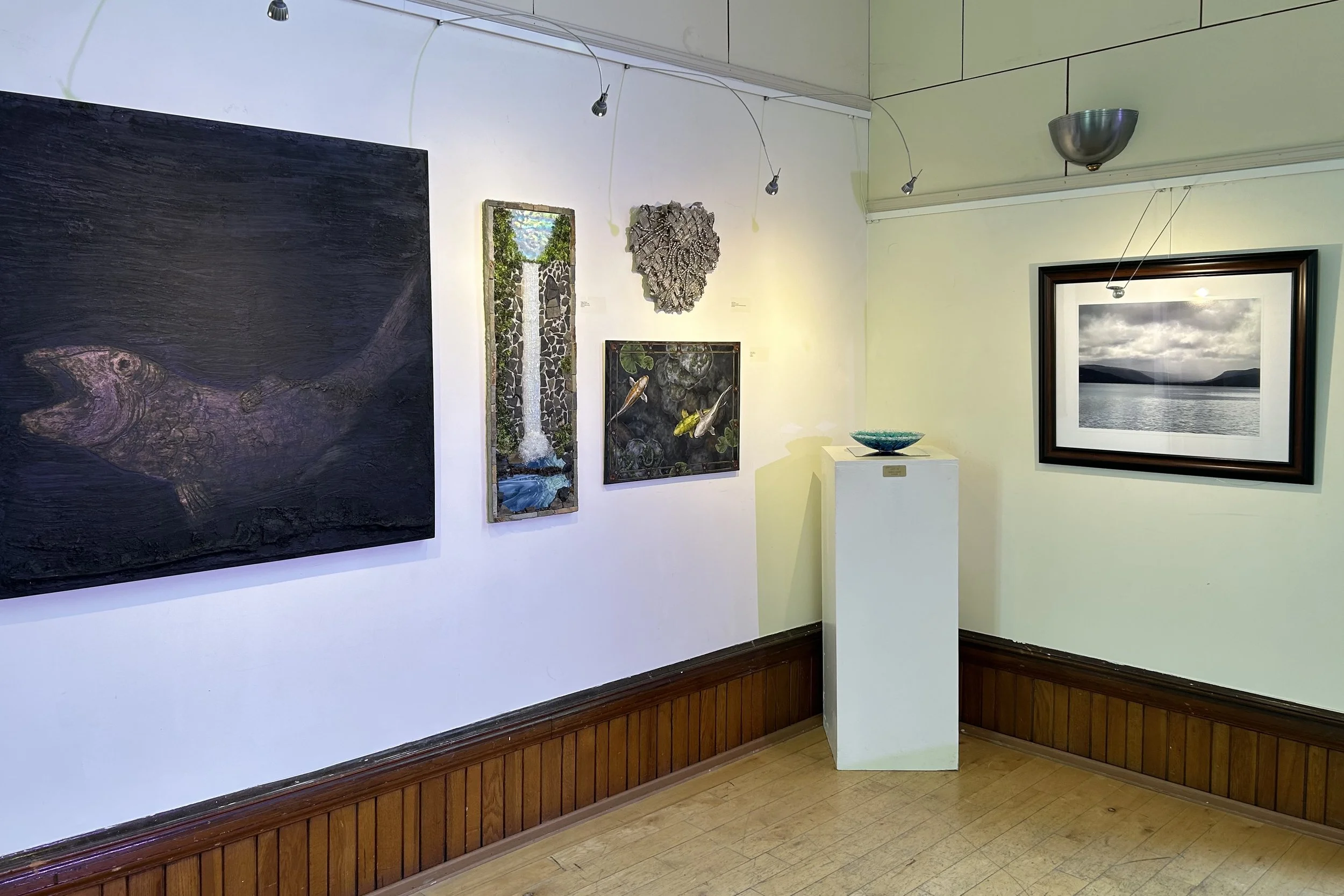 An art gallery wall with various paintings and sculptures, including a water scene with rocks, a metallic floral sculpture, a painting of fish, and a black-and-white landscape photograph, with a white pedestal displaying a decorative bowl and a spur-type light fixture.