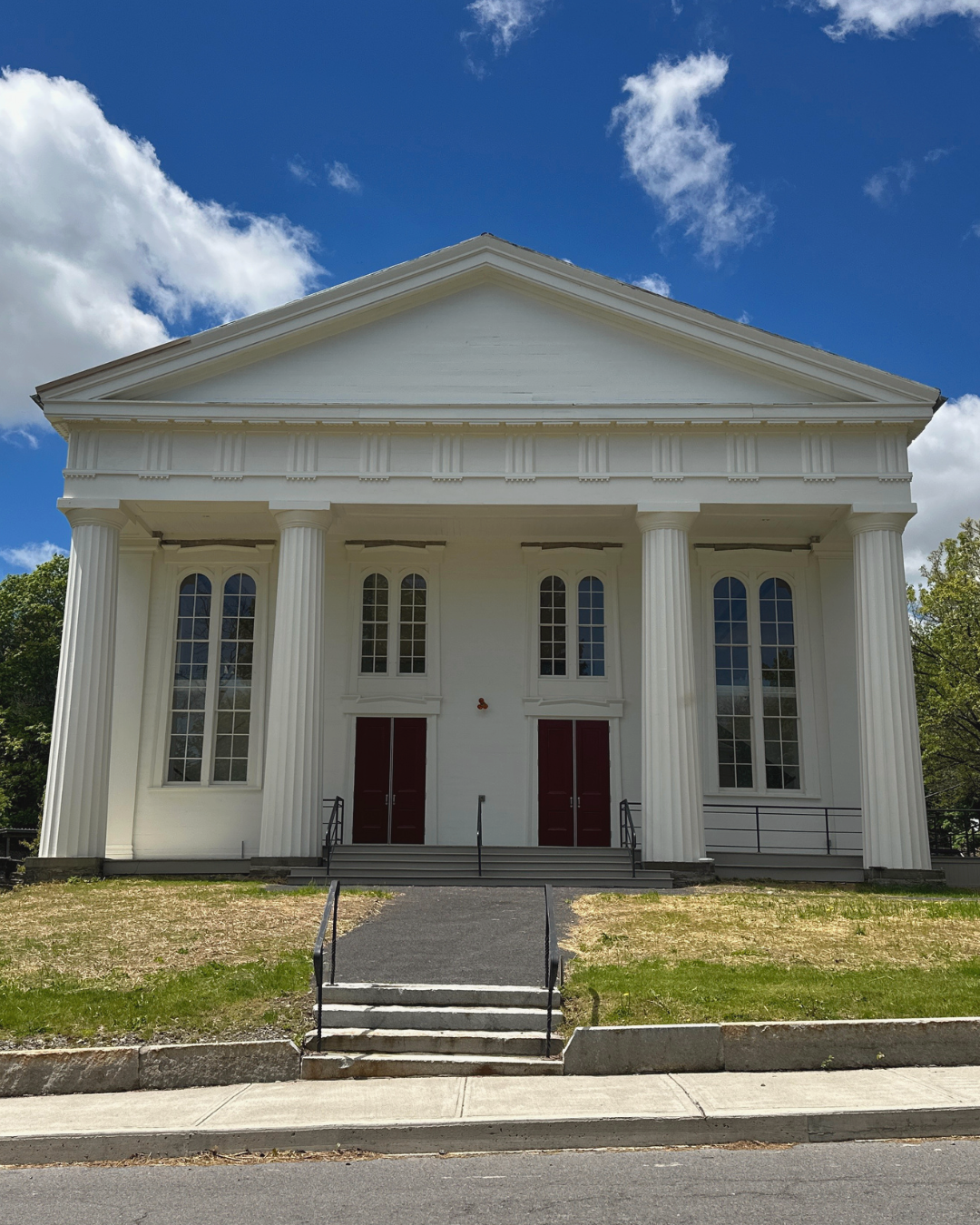 A white neoclassical church with tall columns, red doors, and large arched windows, set against a blue sky with white clouds.