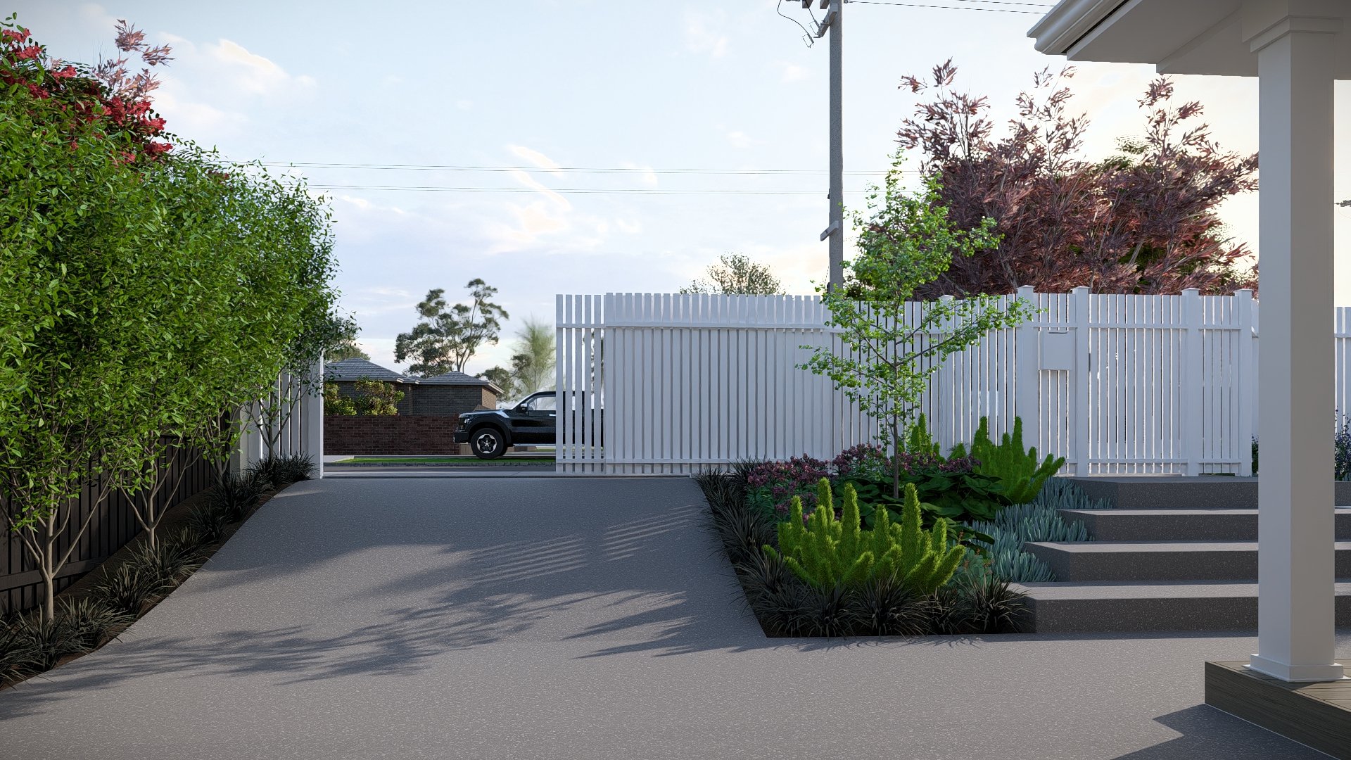 Residential front yard with a concrete driveway, garden beds with shrubs and small trees, white picket fence, concrete steps, and a house porch with a white pillar. A black truck is visible on the street beyond the fence under a partly cloudy sky.