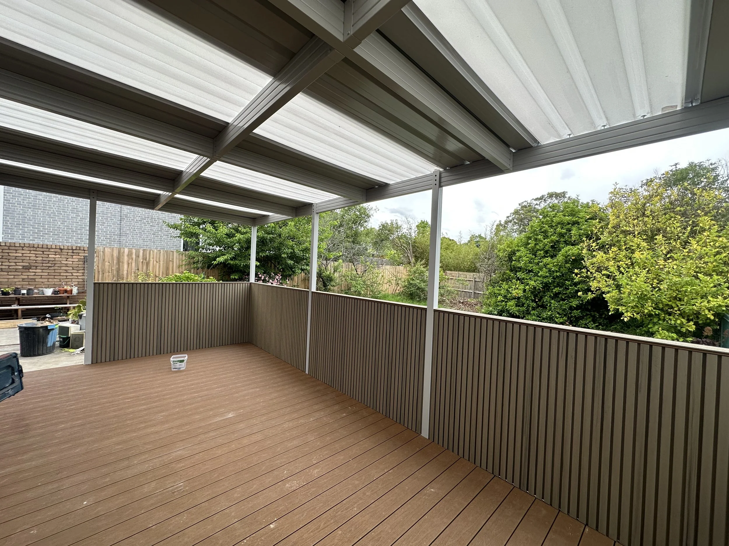 Empty outdoor balcony with wooden flooring, metal railing, and a metal roof, overlooking a backyard with trees and a wooden fence.