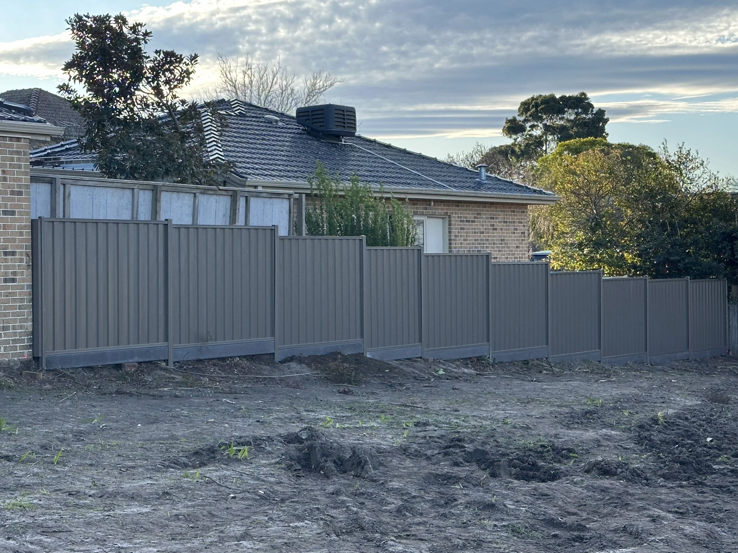 A residential backyard with a brown brick house, a black tiled roof, and a gray metal fence, with trees and sky in the background.