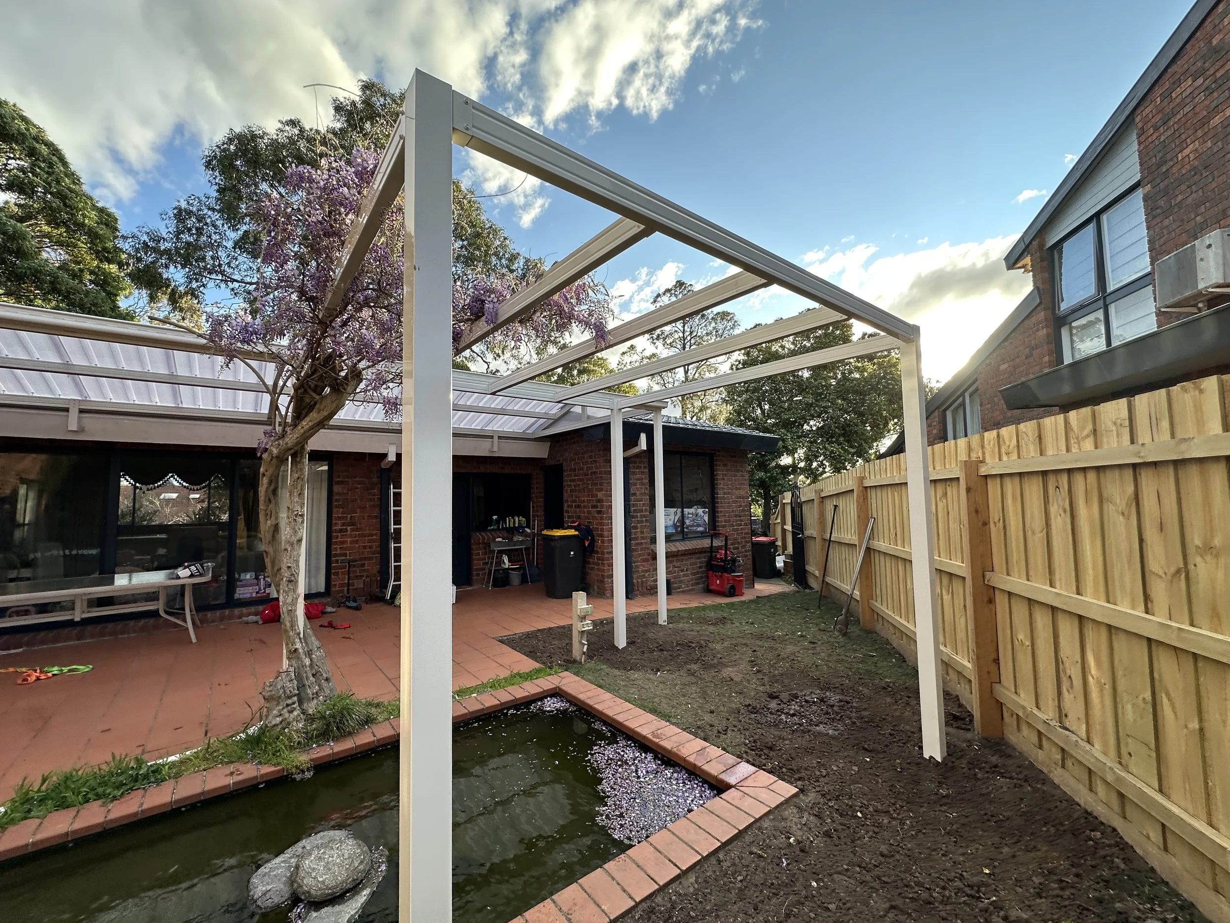 Backyard with a brick house, a small pond with rocks, a wooden fence on the right, a tree with purple flowers, and a white metal frame structure being assembled.