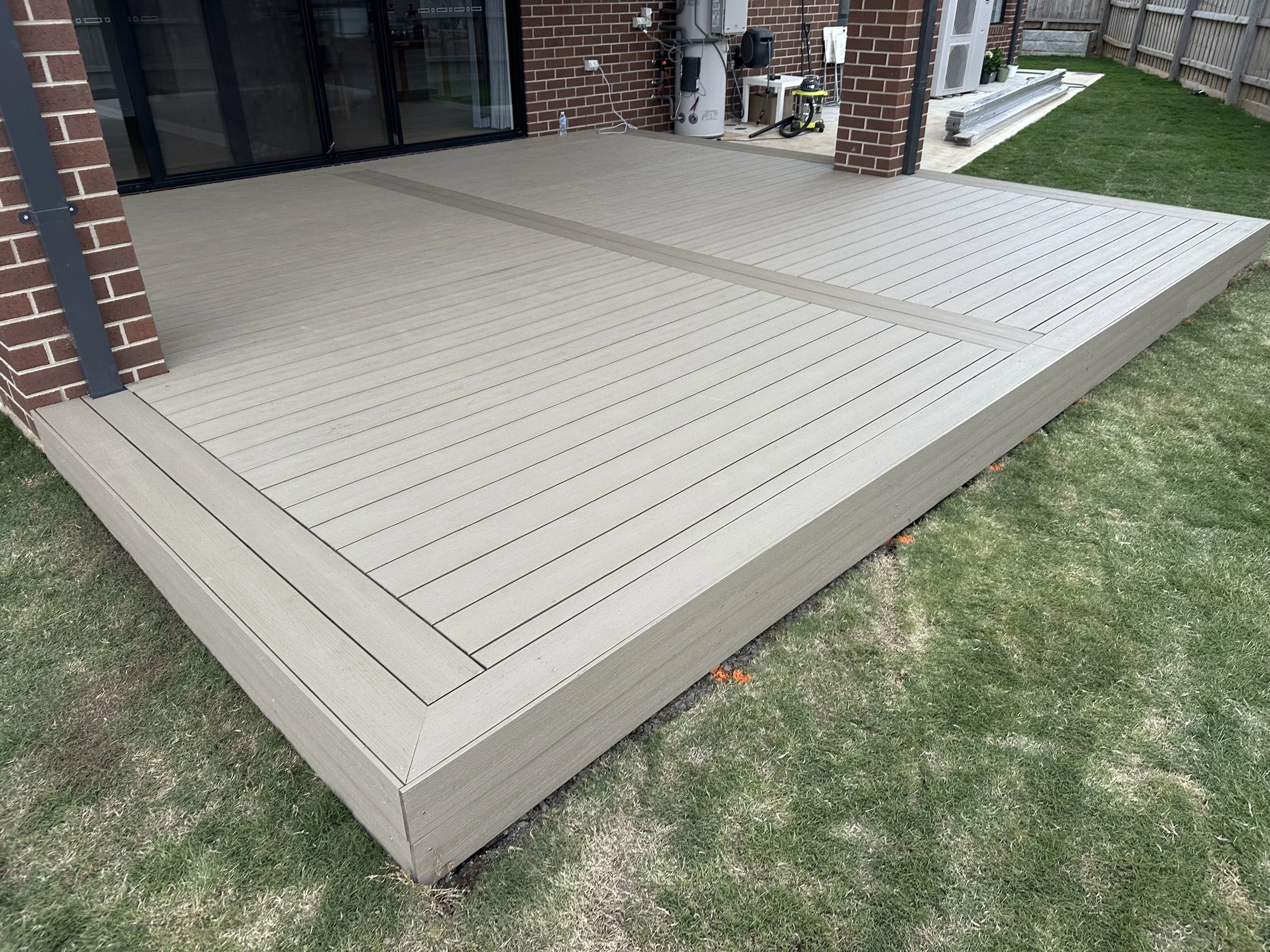Newly built wooden deck attached to a brick house with sliding glass doors, surrounded by a grassy lawn, and with construction materials and tools nearby.