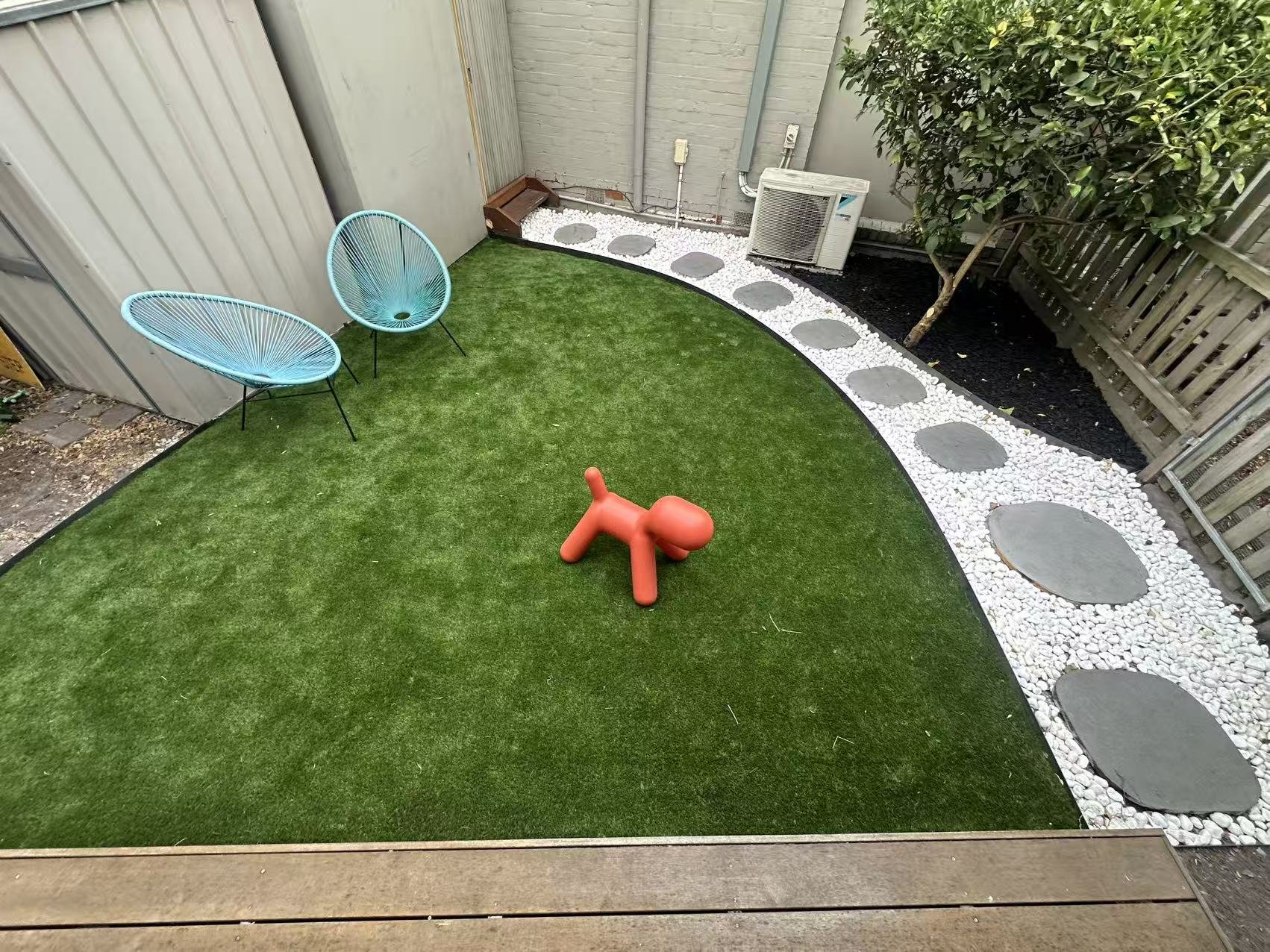 Small backyard with artificial grass, two blue circular chairs, a red dog-shaped toy, white rocks, gray stepping stones, a tree, with a fence on the right and a gray wall on the left, and an air conditioning unit in the corner.
