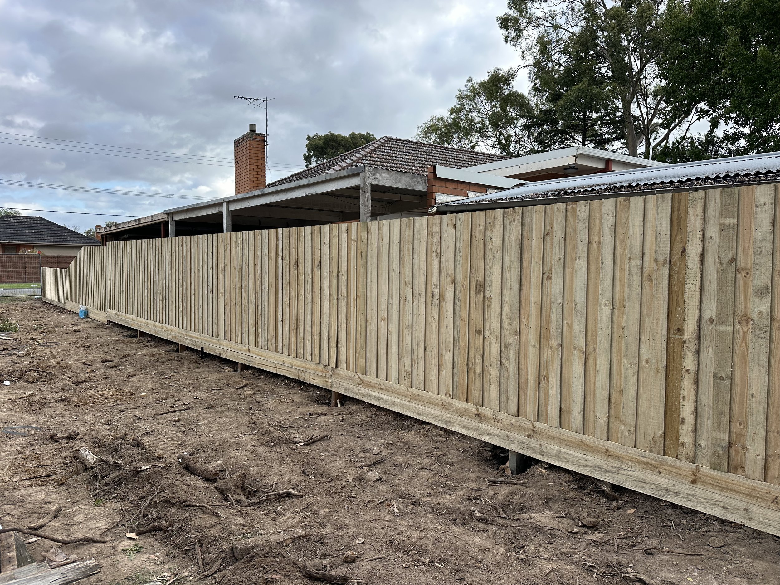 New wooden fence along a backyard with dirt ground, adjacent to a house with brick chimney and roof, under cloudy sky.