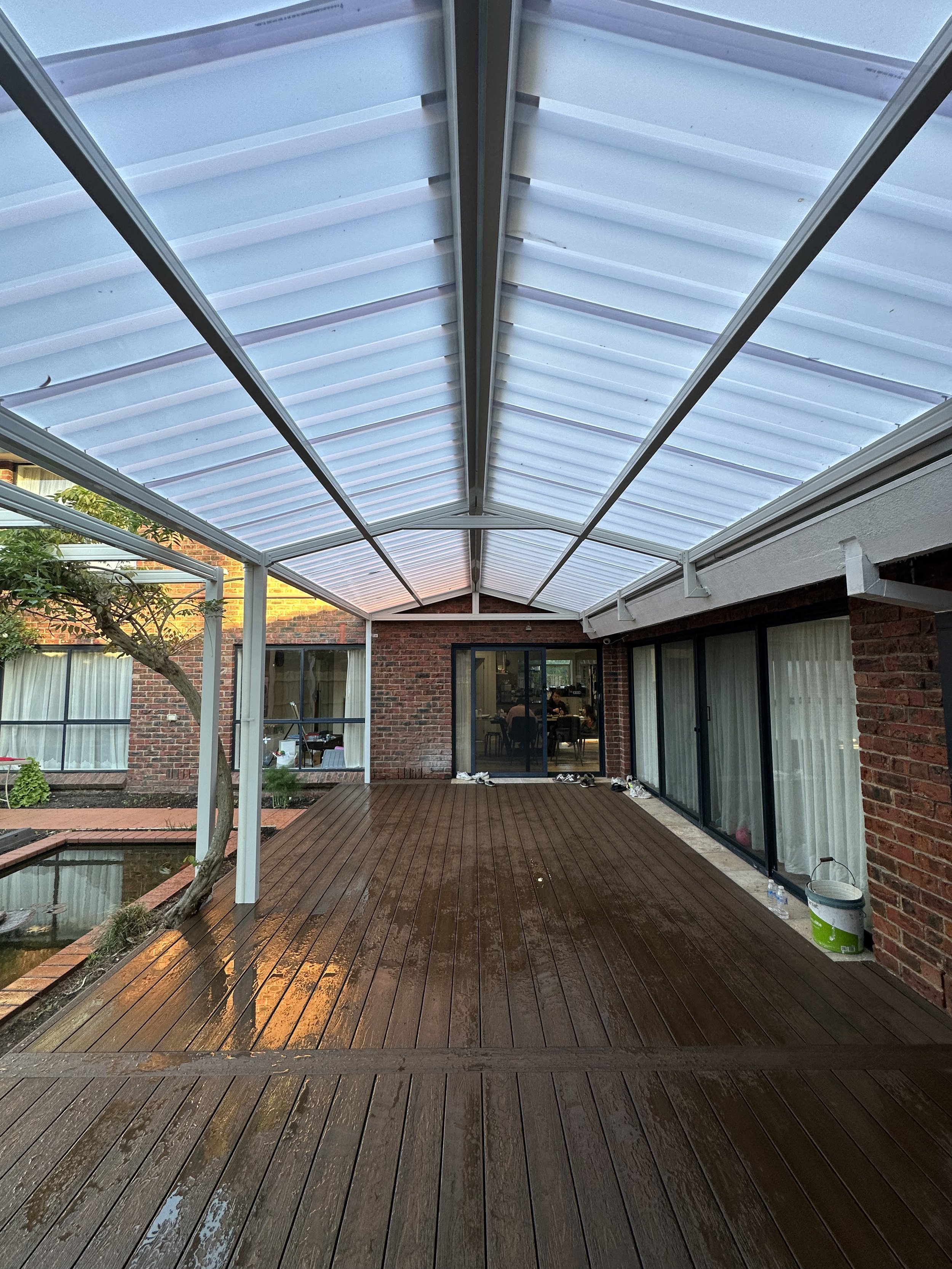 Backyard covered patio with wooden deck, brick house, sliding glass doors, and a transparent roof structure.