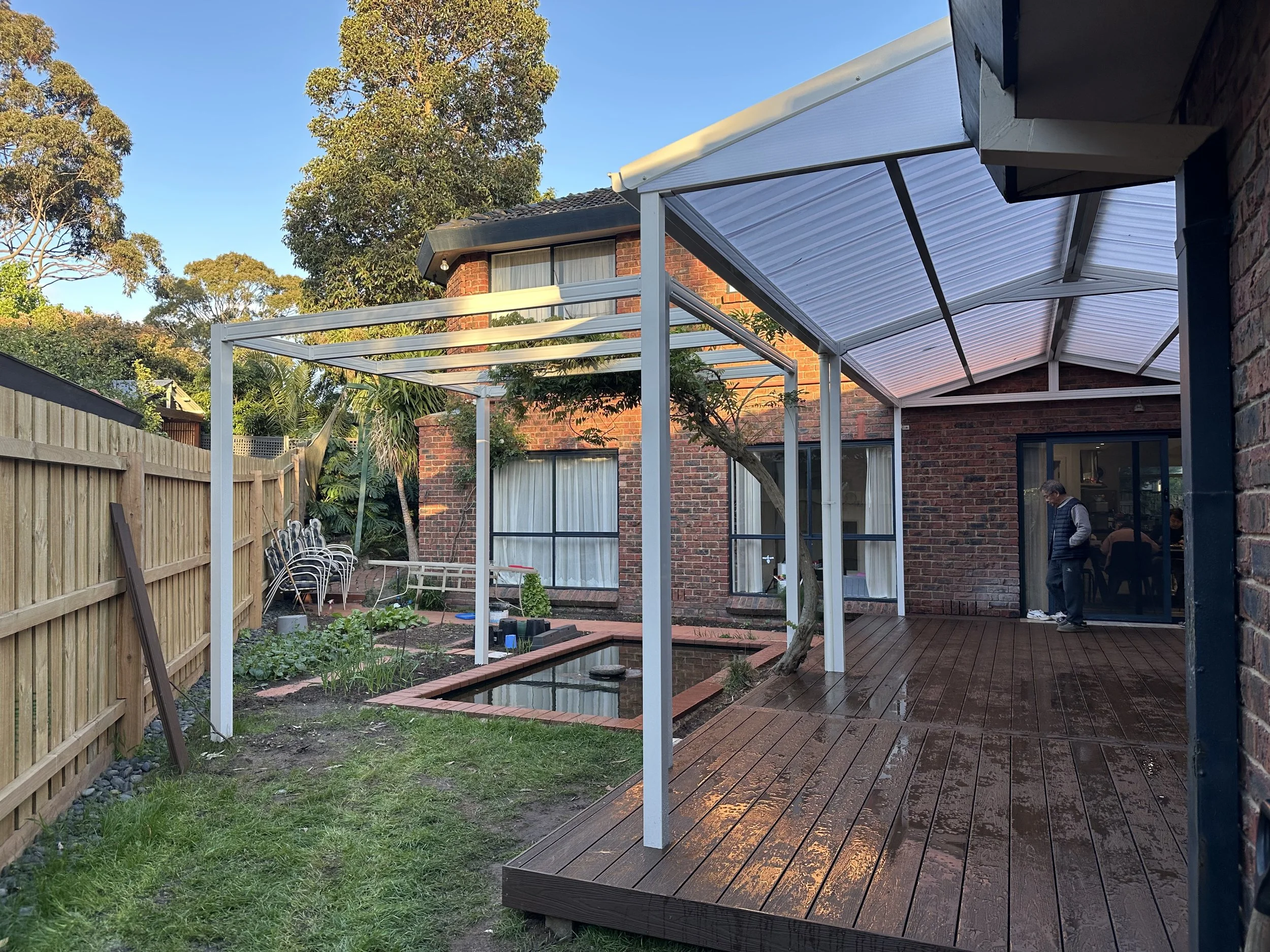A backyard with a small pond, vegetable garden, and wooden deck. There is a covered patio area with a man standing near the sliding glass door of a brick house and trees in the background.