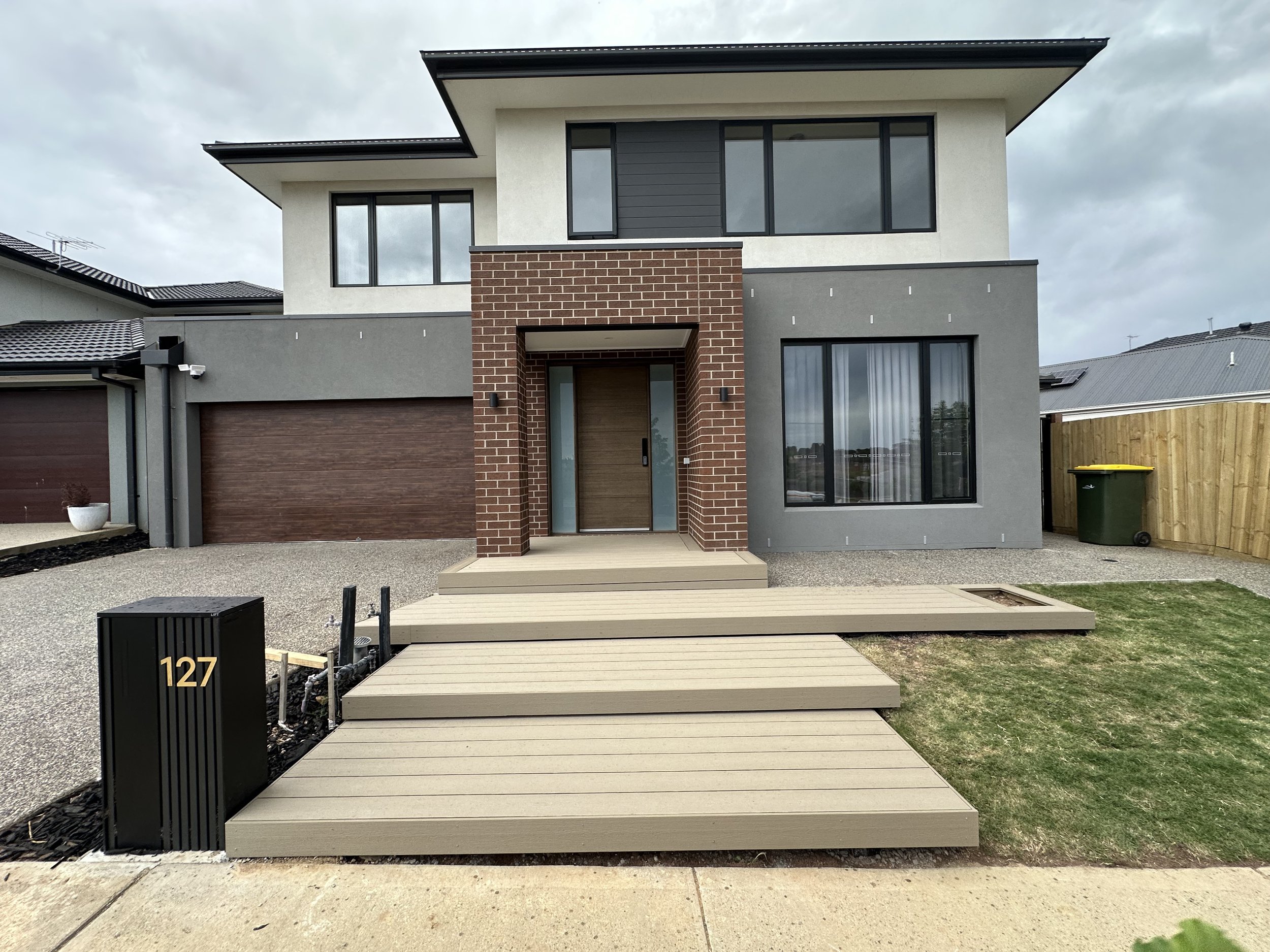 Modern two-story house with a front porch, brick and gray exterior, large windows, and a wooden staircase leading to the front door. House number 127 on a black mailbox in the yard.