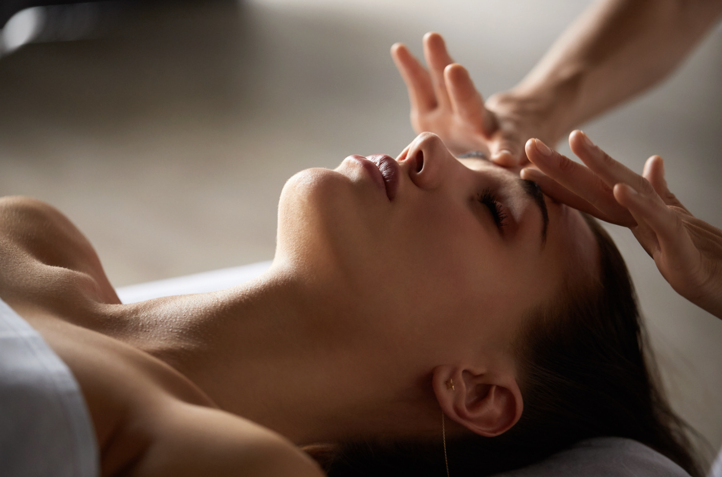 A woman lying down with her eyes closed, receiving a massage on her forehead from a massage therapist.