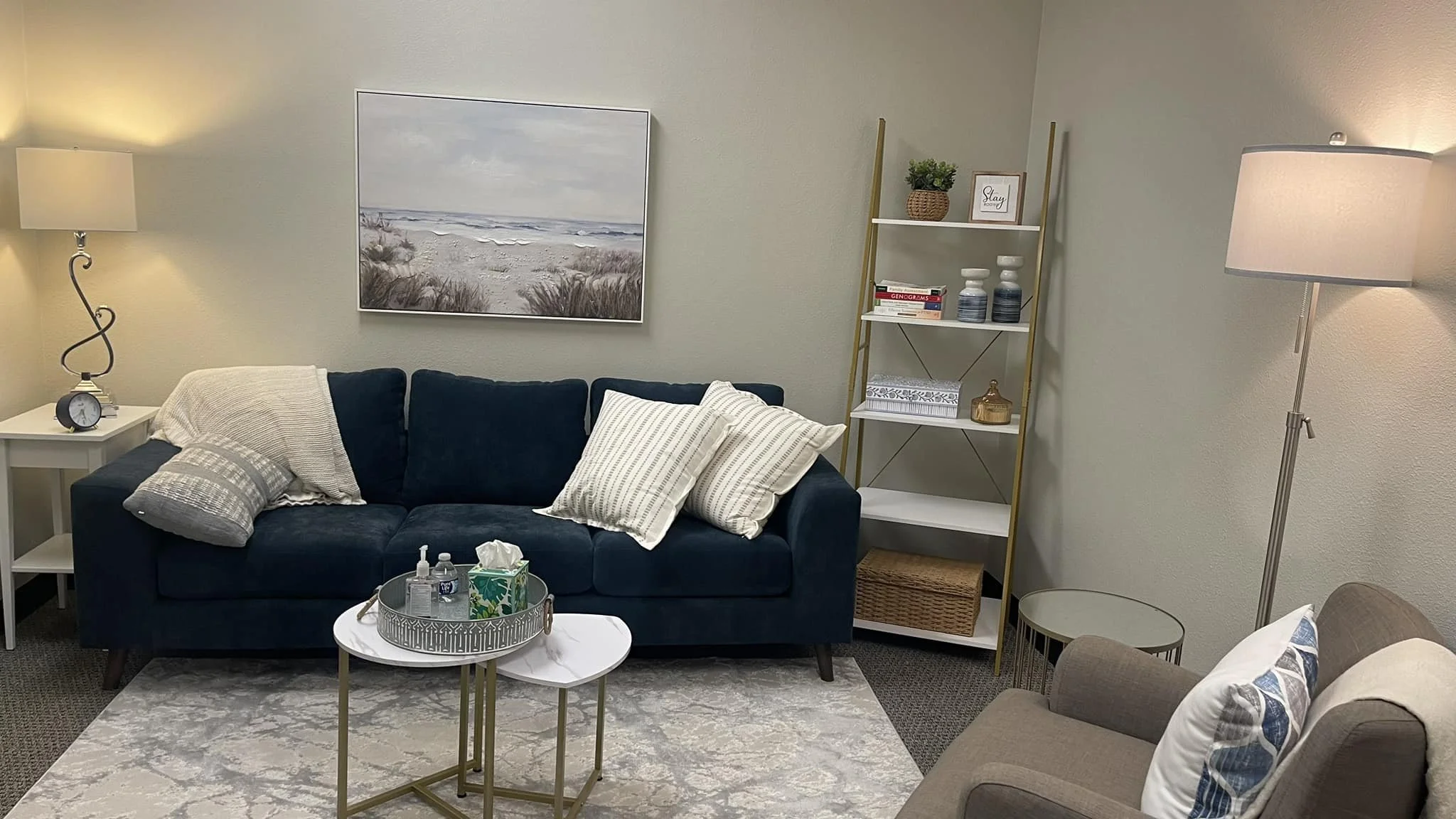 Living room with navy sofa, beige armchair, two side tables, bookshelf, painting of a beach scene, and decorative pillows and lamps.