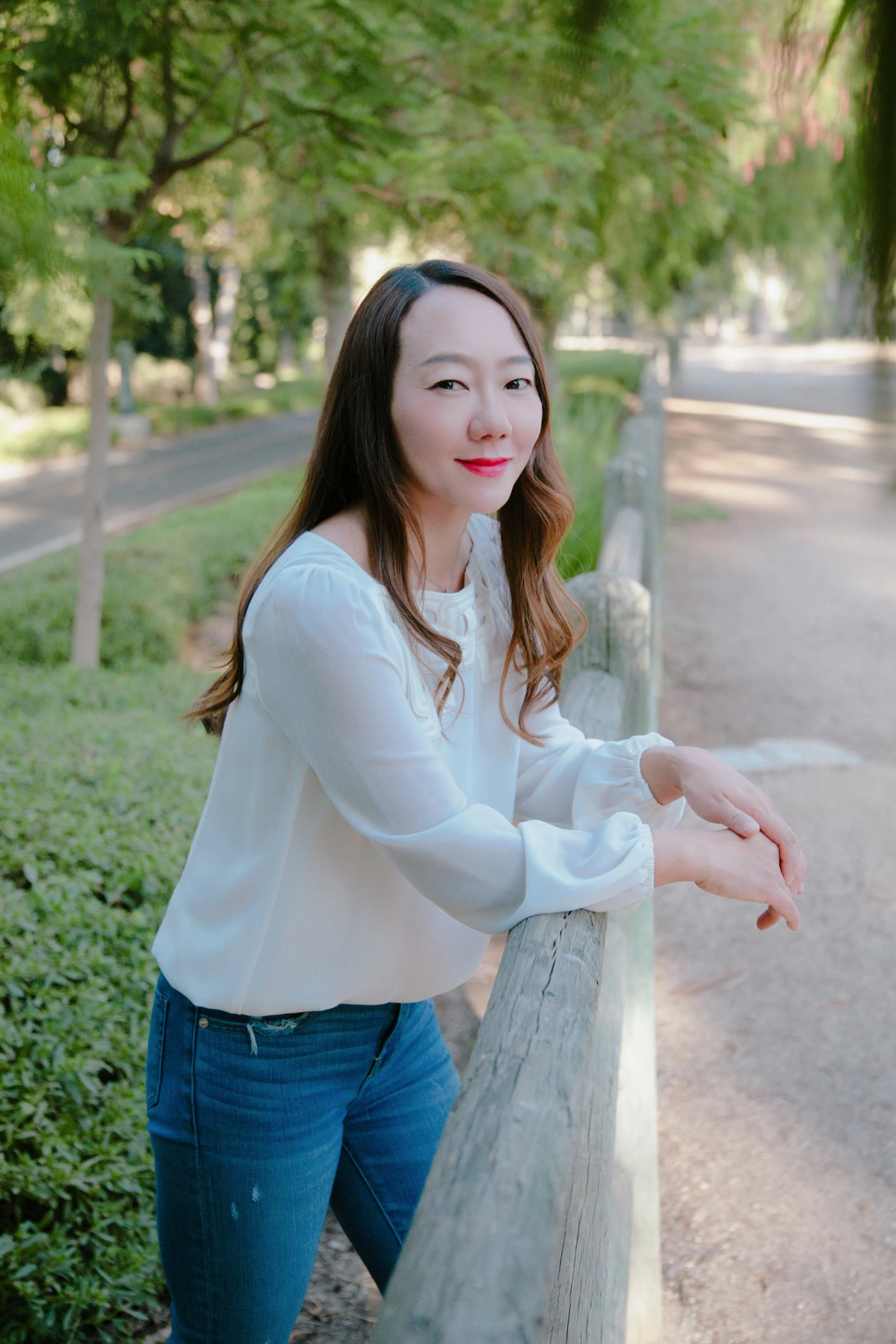 A woman leaning on a wooden fence in a park, smiling at the camera, with trees and greenery in the background.