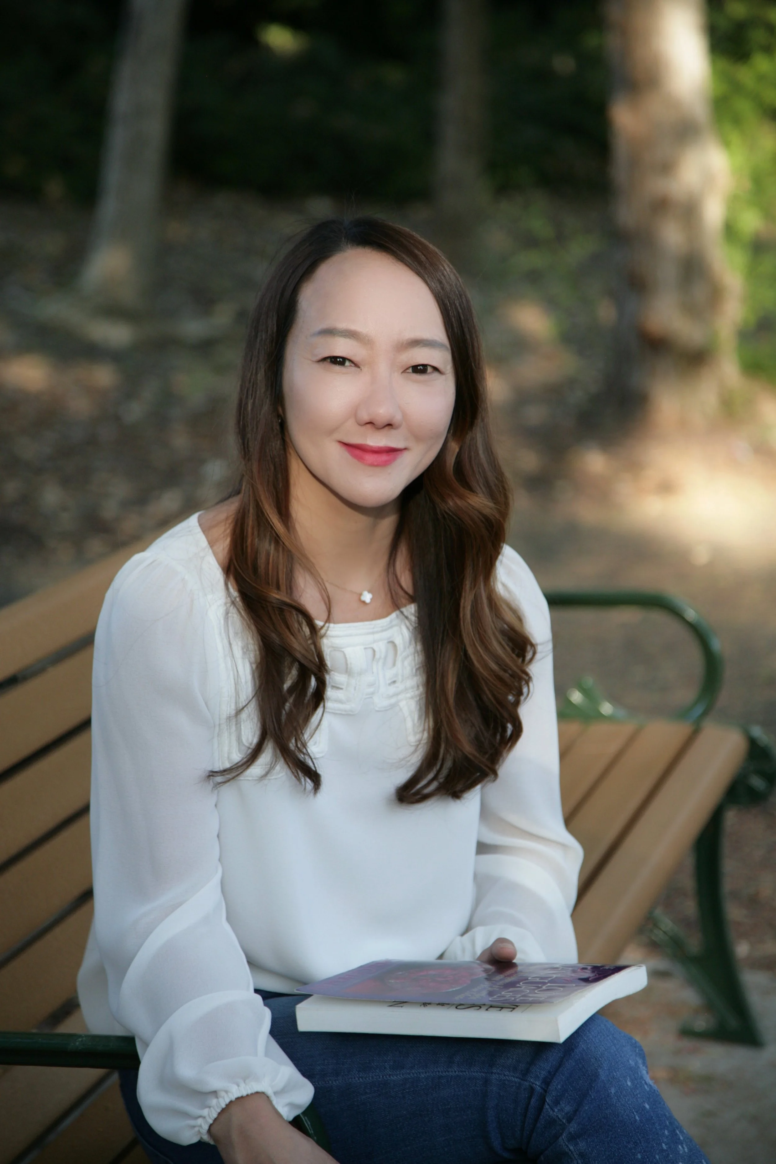 A woman with long wavy brown hair, wearing a white blouse, sitting on a park bench in a wooded area, holding a book.