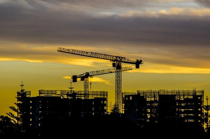 Silhouette of construction cranes and building frameworks against a yellow and gray cloudy sky at sunset.