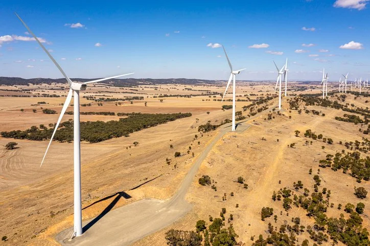 An aerial view of a wind farm with multiple large wind turbines on a dry, open landscape under a blue sky with scattered clouds.