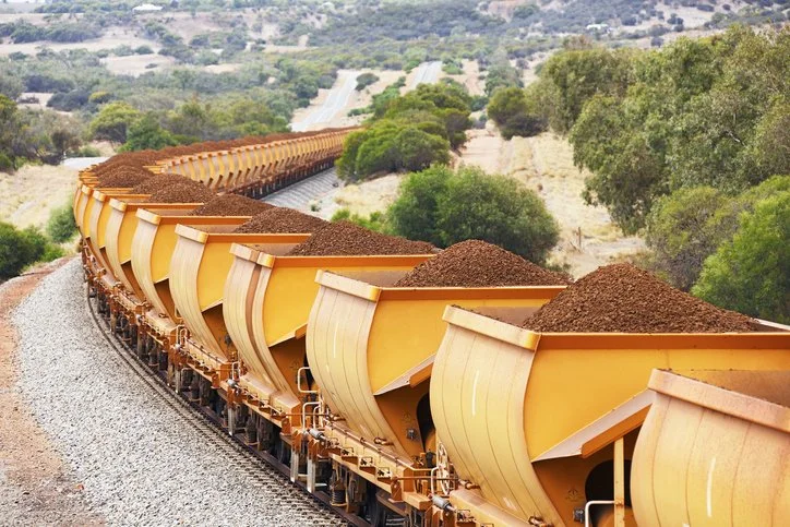A freight train carrying brown soil on yellow hopper cars moving through a rural area with green trees and rolling hills.