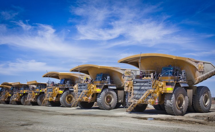 A row of large yellow mining dump trucks parked on a dirt lot under a partly cloudy blue sky.