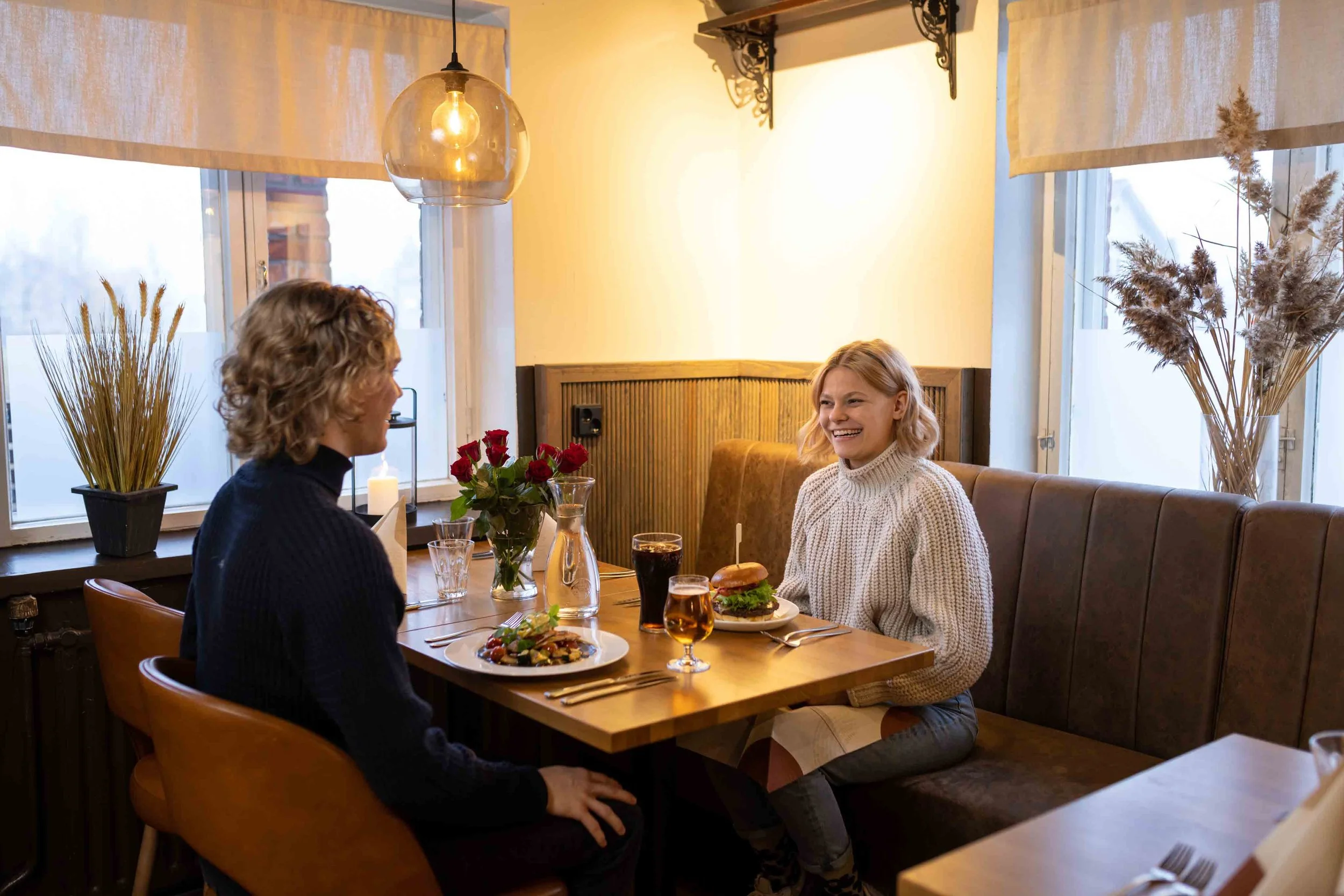 Kaksi ihmistä nauttimassa ateriaa ravintolassa, keskustellen ja nauraen, pöydällä ruoka, juomia ja kukkakimppu.