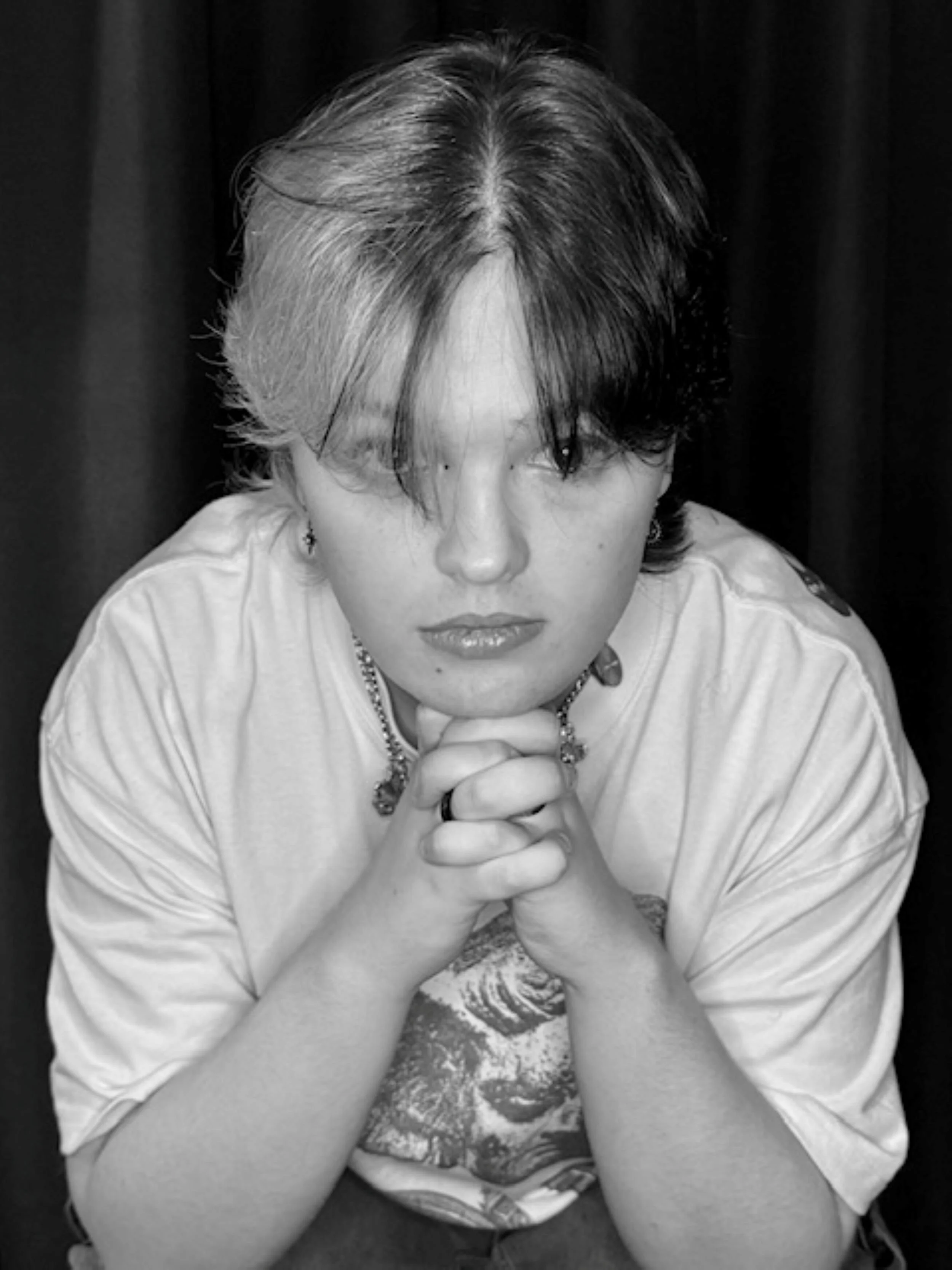 Black and white photo of a person with short two-tone hair, wearing jewelry, leaning forward with hands clasped under chin, dark background.