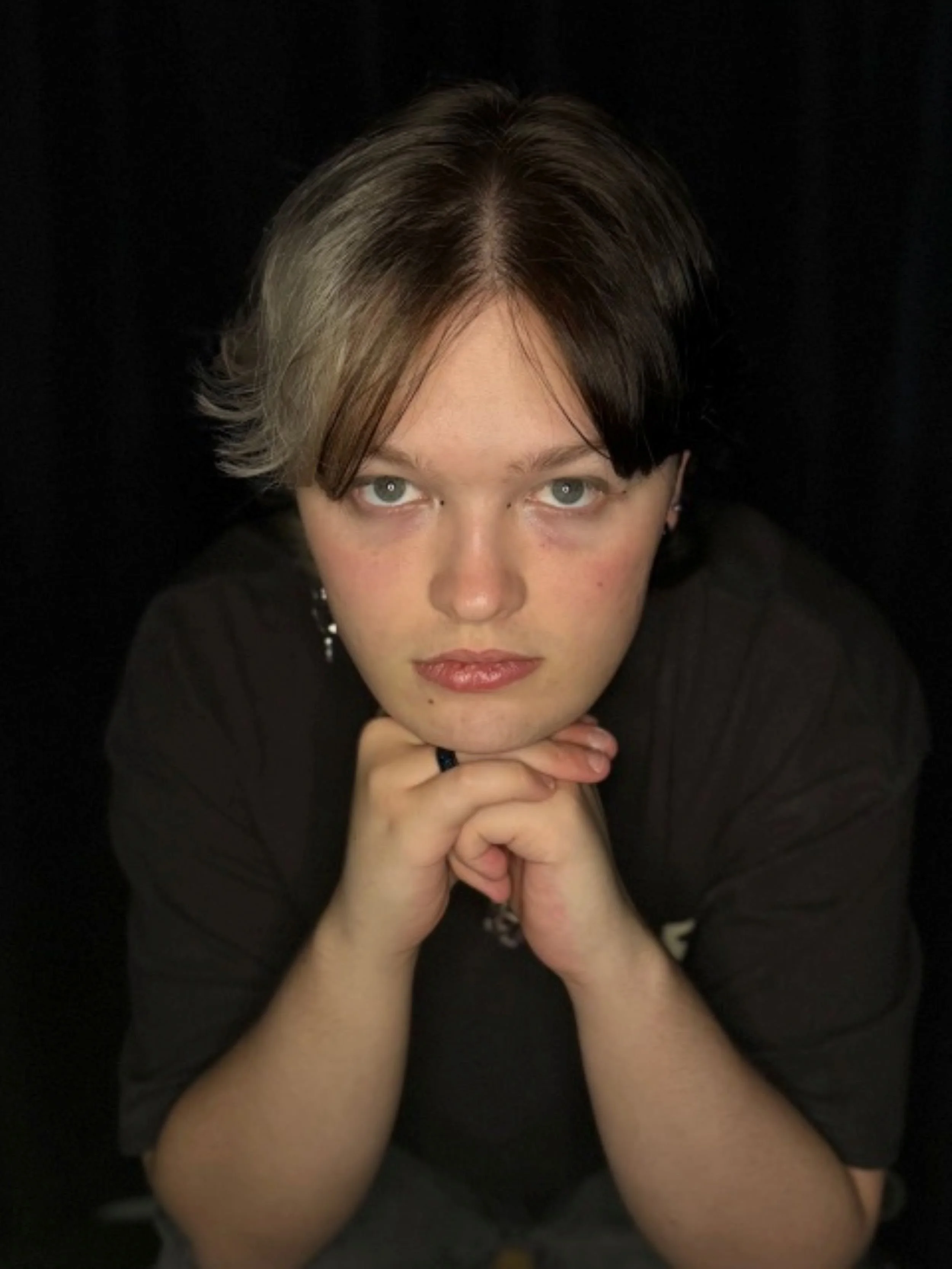Close-up portrait of a person with short, two-toned hair, resting their chin on clasped hands, wearing a black shirt, against a dark background.