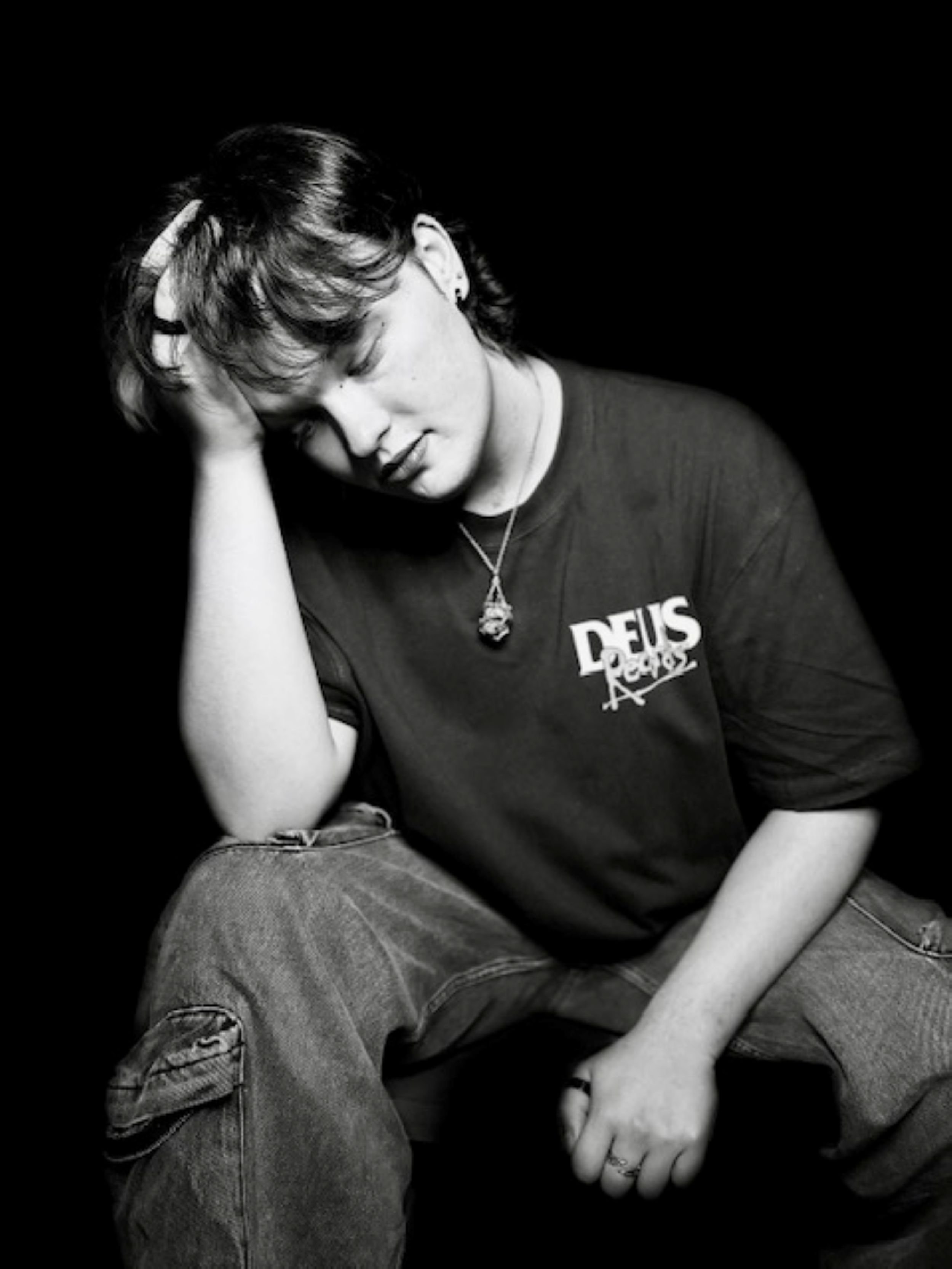 Black and white photo of a person sitting with their head tilted, hand on their head, wearing a t-shirt with text and a necklace, against a dark background.