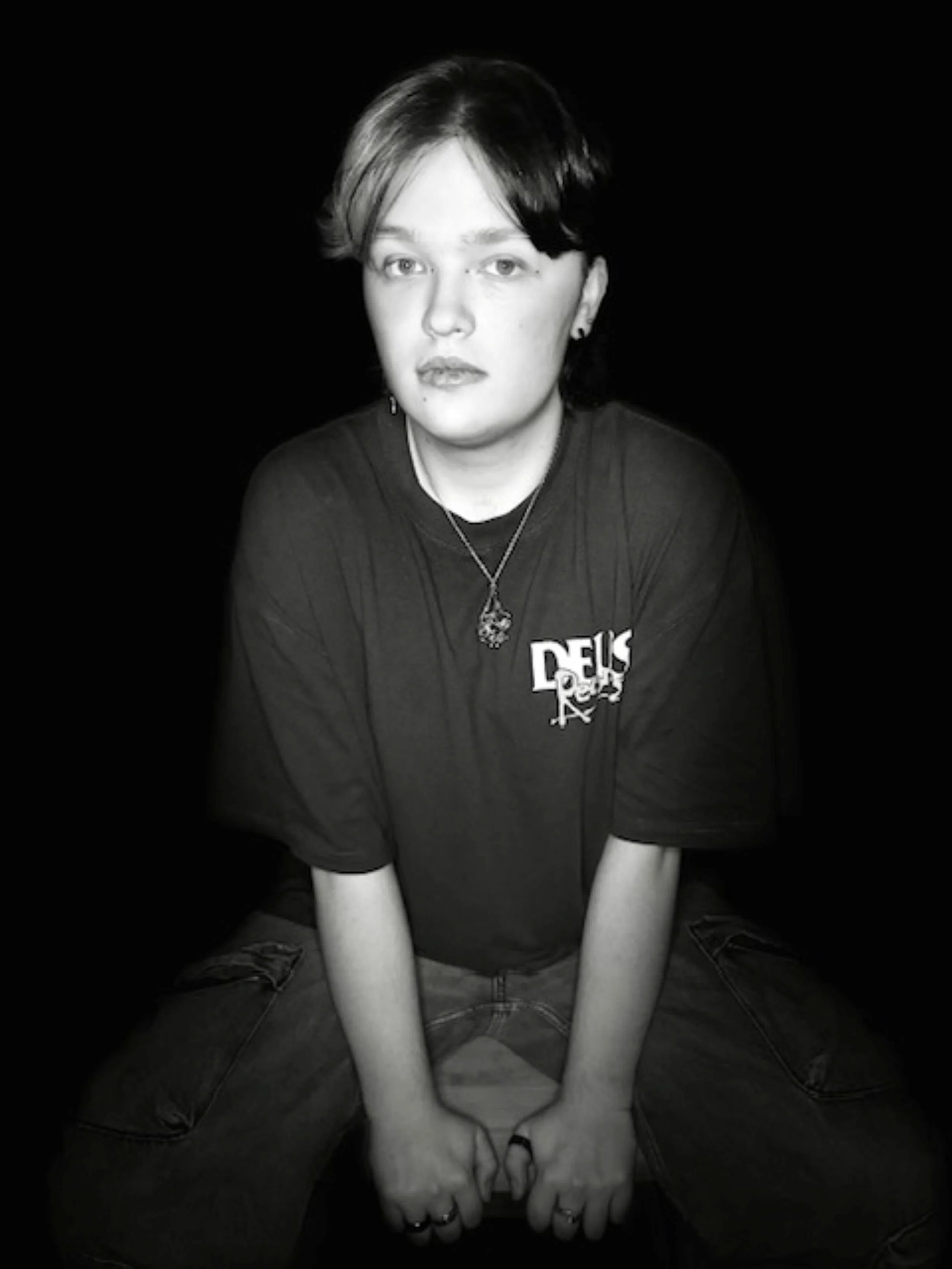 Black and white photo of a person with short hair, wearing a dark t-shirt and jewelry, sitting against a black background.