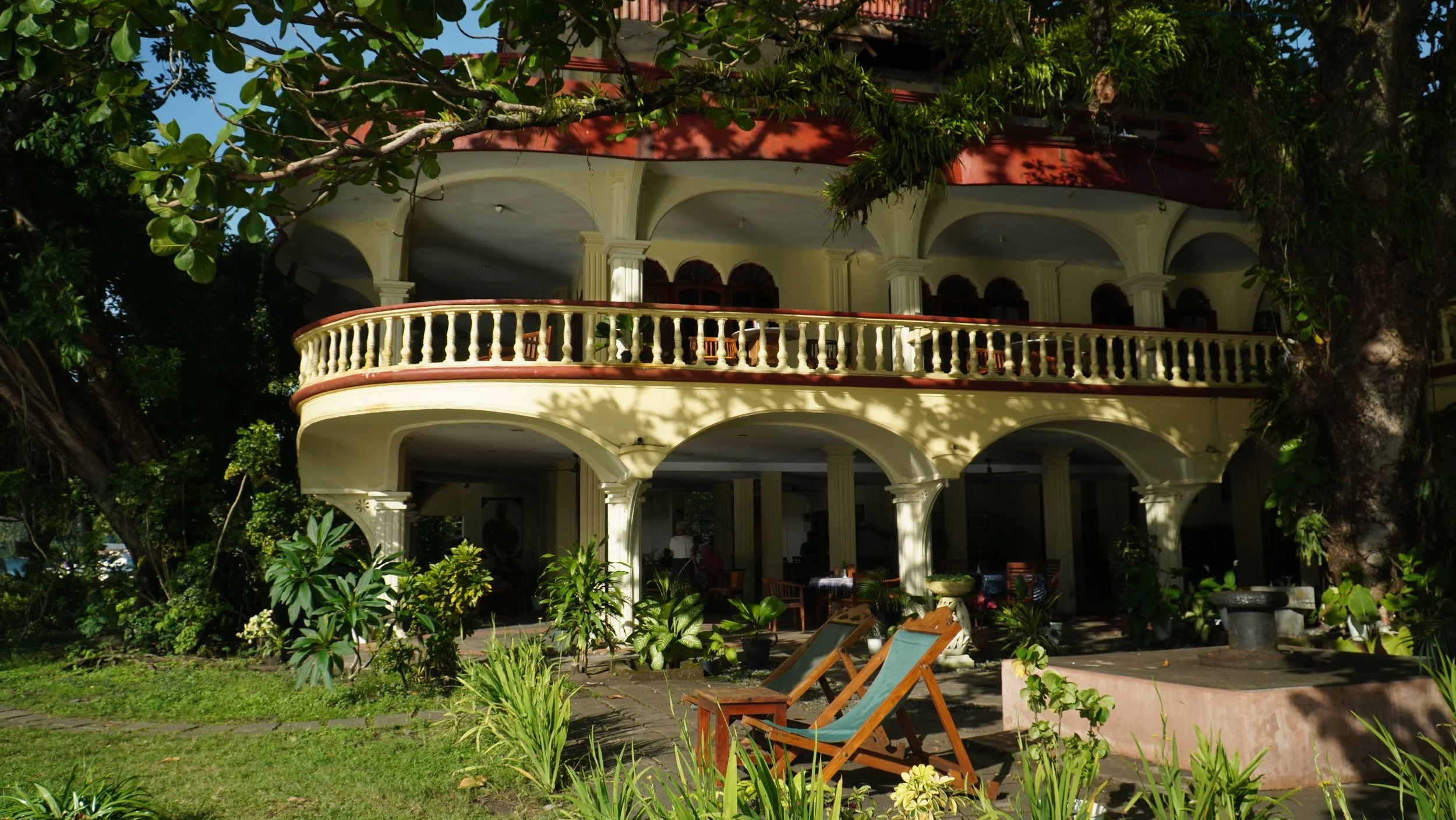 A multi-story house with a rounded balcony, painted in yellow with white columns, partially obscured by trees and lush green plants in the foreground.