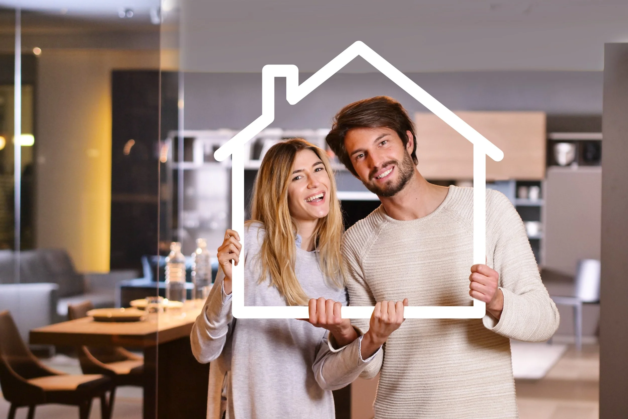 A smiling couple holding a house-shaped frame, smiling and looking at the camera in a modern kitchen or dining area.