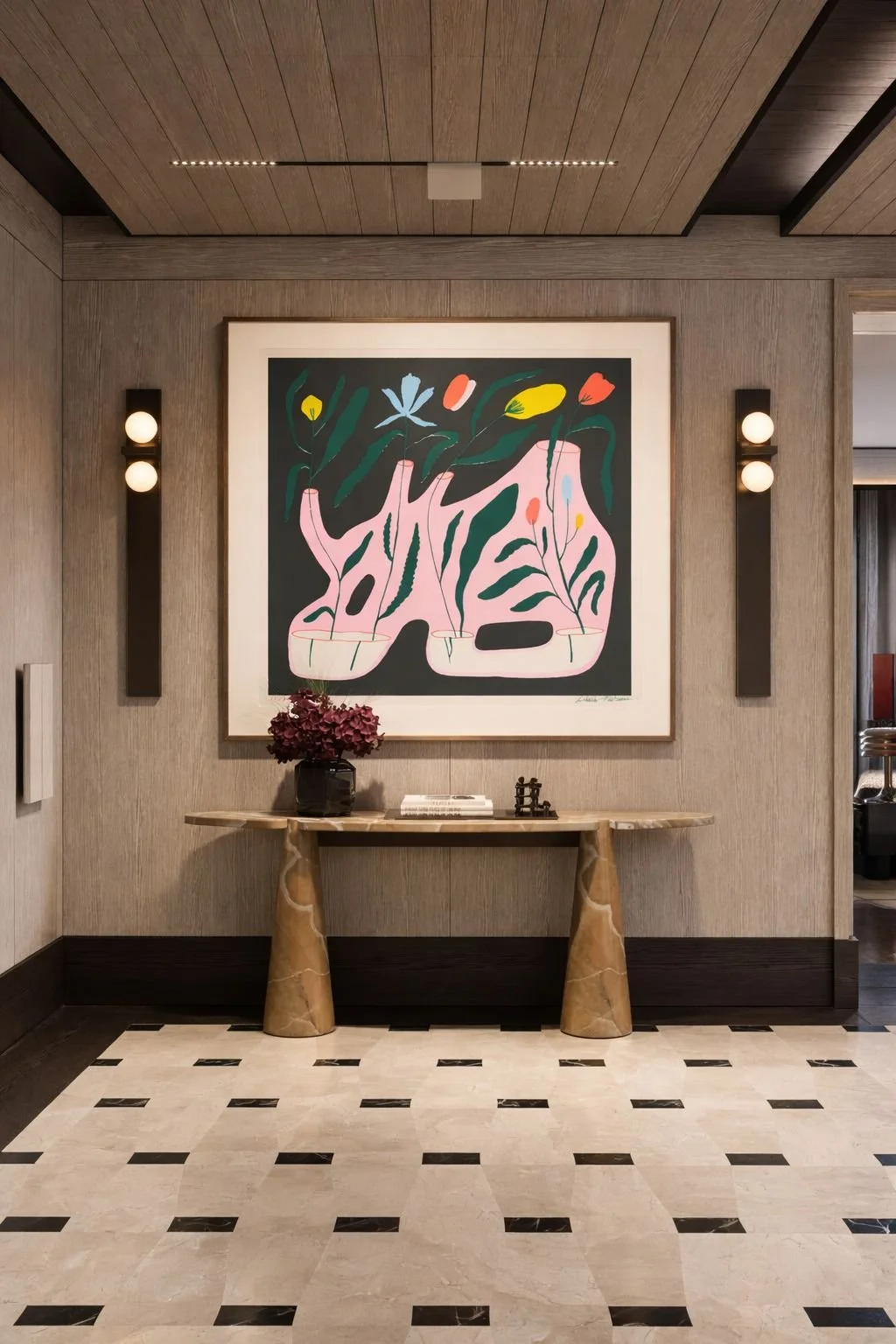 Interior view of a stylish room with a wooden ceiling, beige walls, and a black and white checkered marble floor. A large abstract floral artwork hangs on the wall, flanked by two black wall sconces with round white lights. Below the artwork, a light-colored wooden console table holds a dark vase with purple flowers, a small stack of books, and decorative items.