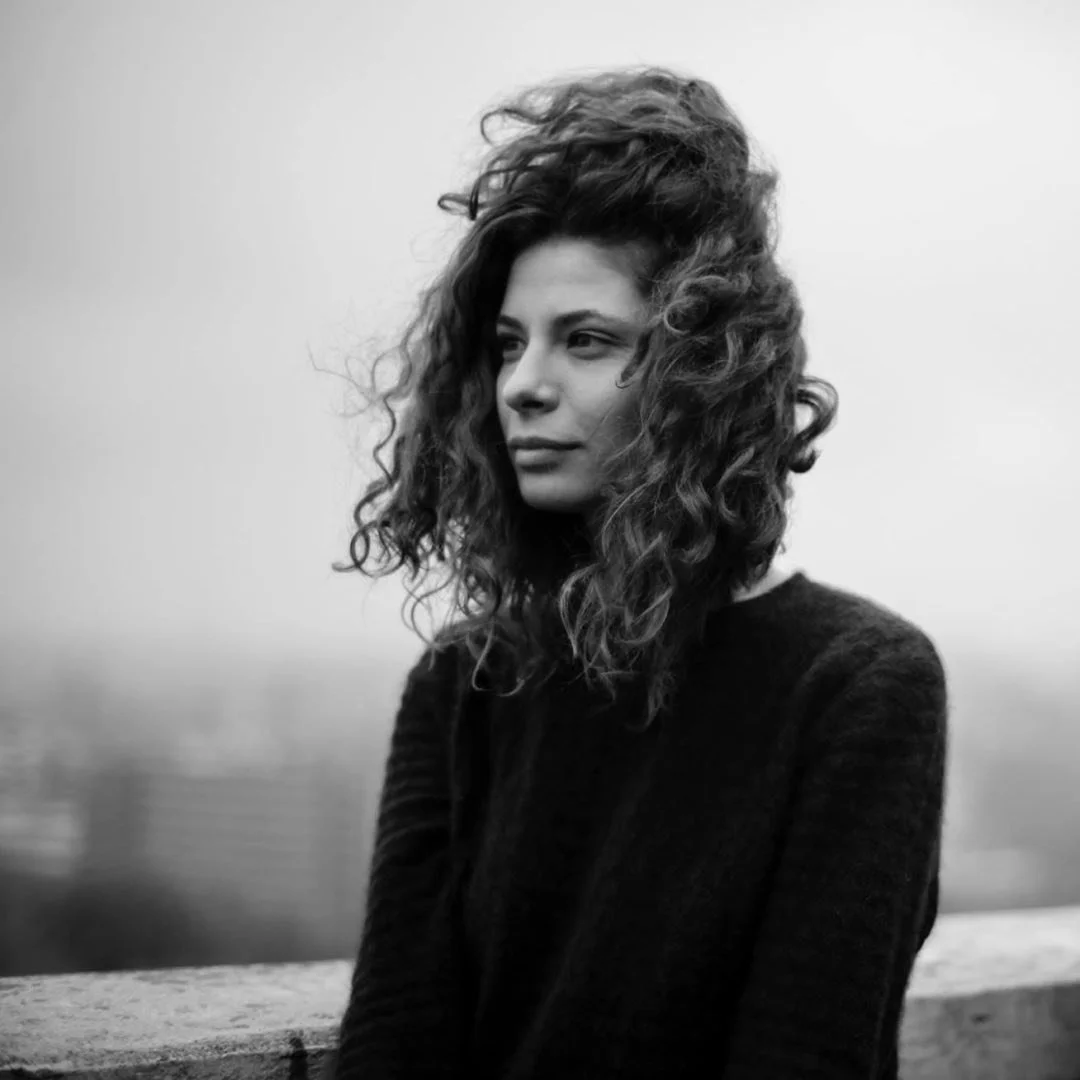 Black and white photo of a woman with curly hair standing outdoors by a railing, looking to the side with a contemplative expression.