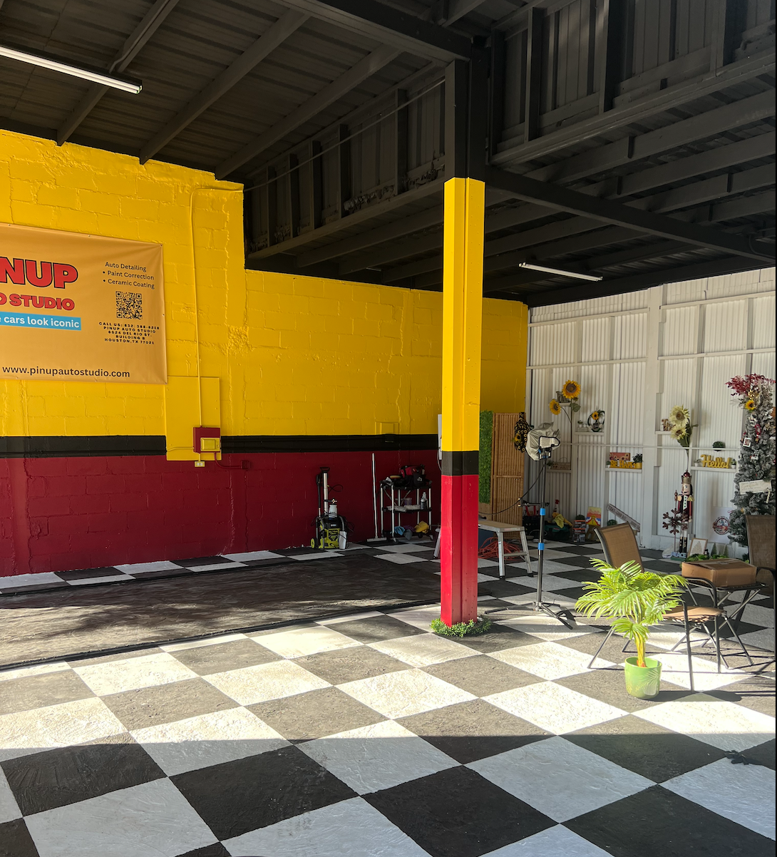 Indoor space with black and white checkered flooring, yellow painted brick wall with a red lower section, a yellow pillar with red and black accents, and various decorations including flowers and a small potted plant.