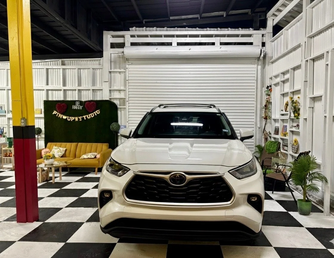 A white Toyota SUV parked inside a room with a black-and-white checkered floor, decorated with a yellow couch, shelves with plants, and a green wall backdrop with hearts and flowers.