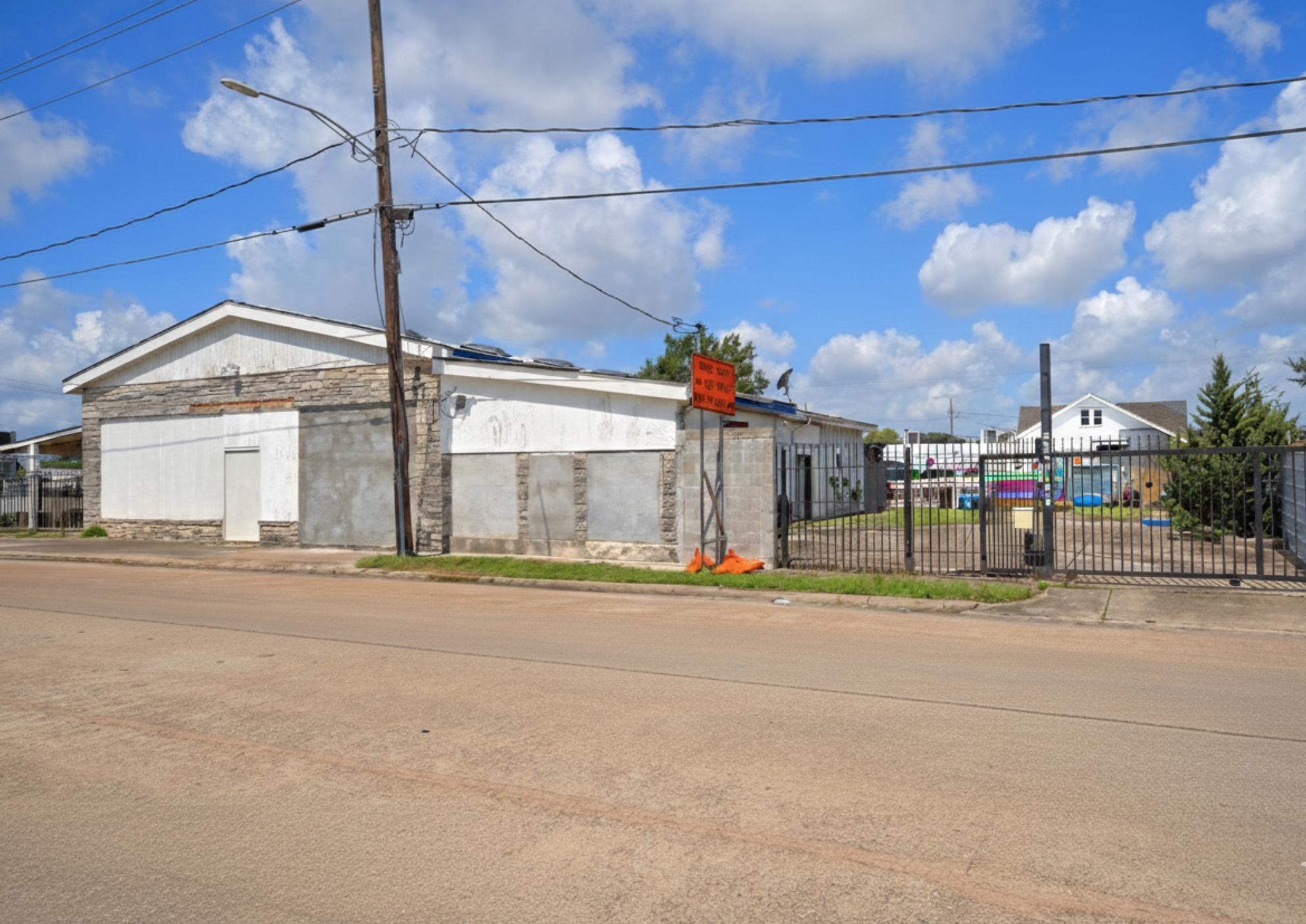 A building with boarded-up windows and a sign that says 'Street Closed' in front of a fenced yard with colorful kayaks and a small house in the background. Clear blue sky with clouds.