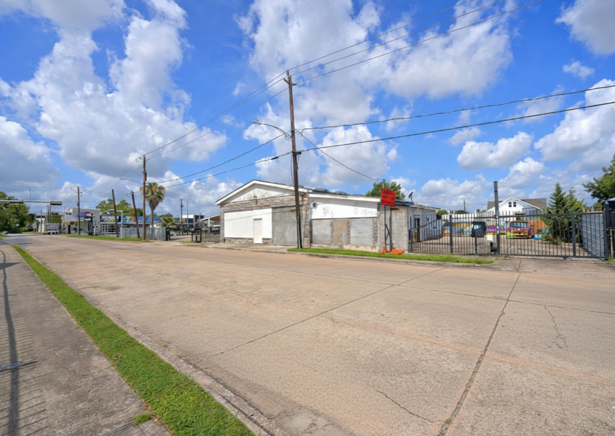 Street view with a white building, utility poles, a parking lot with cars, and a blue sky with scattered clouds.
