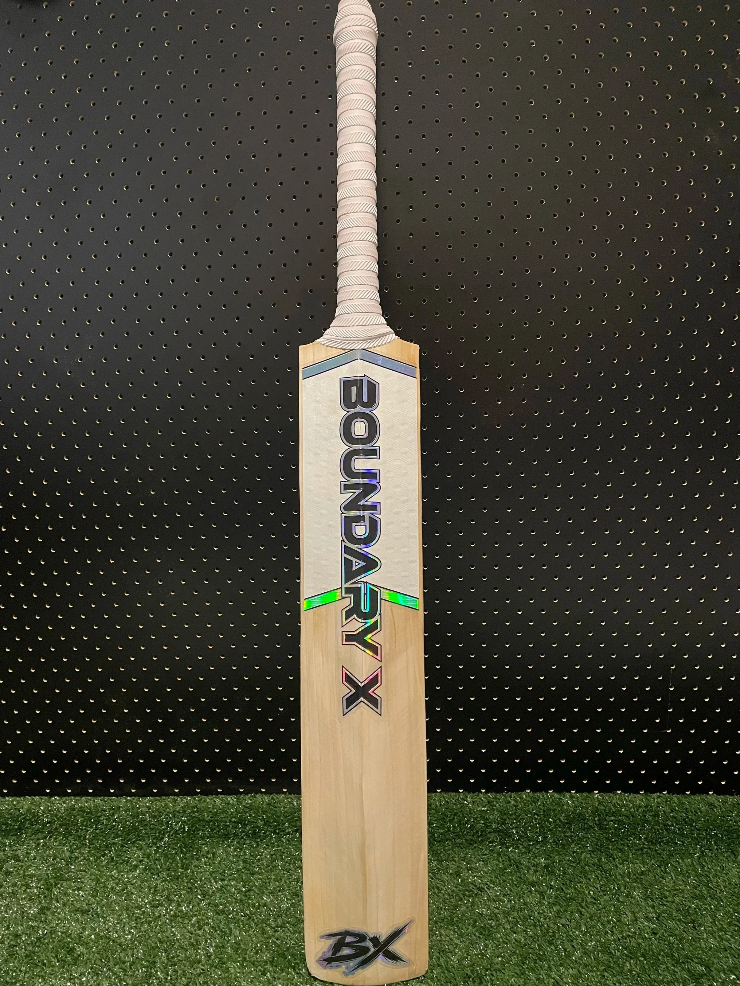 A cricket bat with the words 'BOUNDARY X' written on the front, standing upright on a green surface against a black perforated wall background.