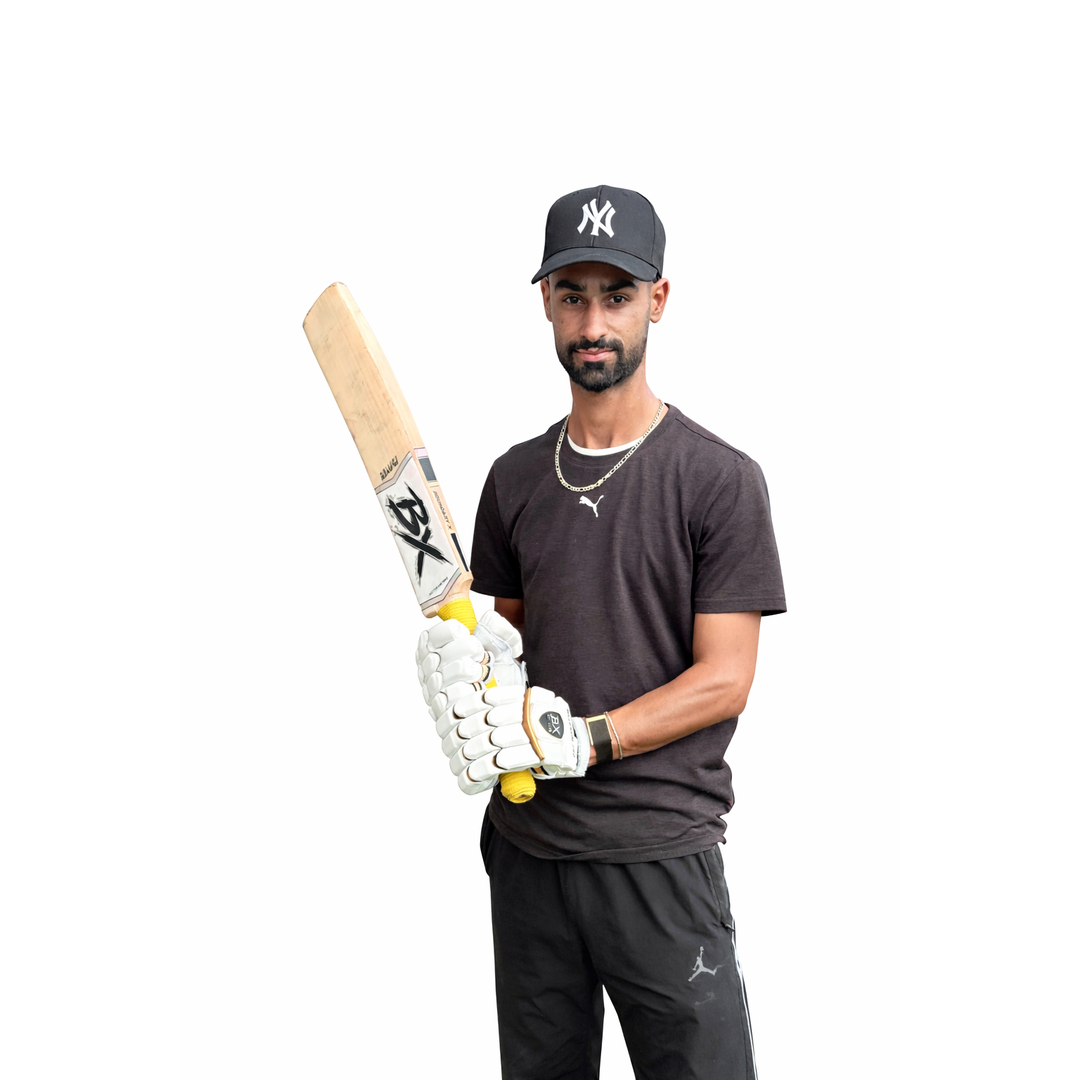 Man wearing a black NY Yankees cap, black T-shirt, and black shorts holding a cricket bat and wearing batting gloves, standing against a white background.
