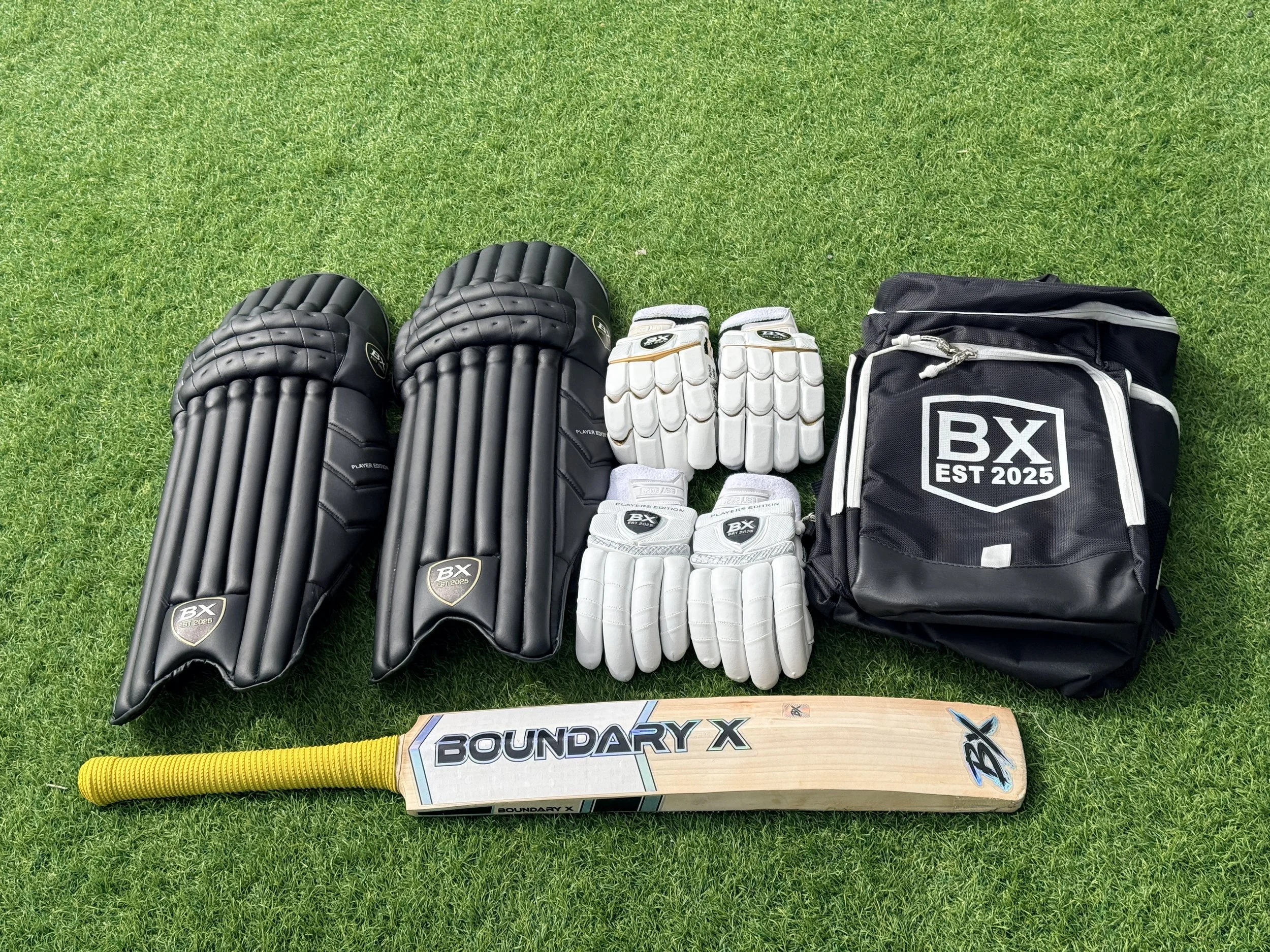 Cricket gear including two black leg guards, four pairs of white batting gloves, a black backpack with 'BX EST 2025' logo, and a cricket bat with yellow handle and 'Boundary X' label, laid out on grass.