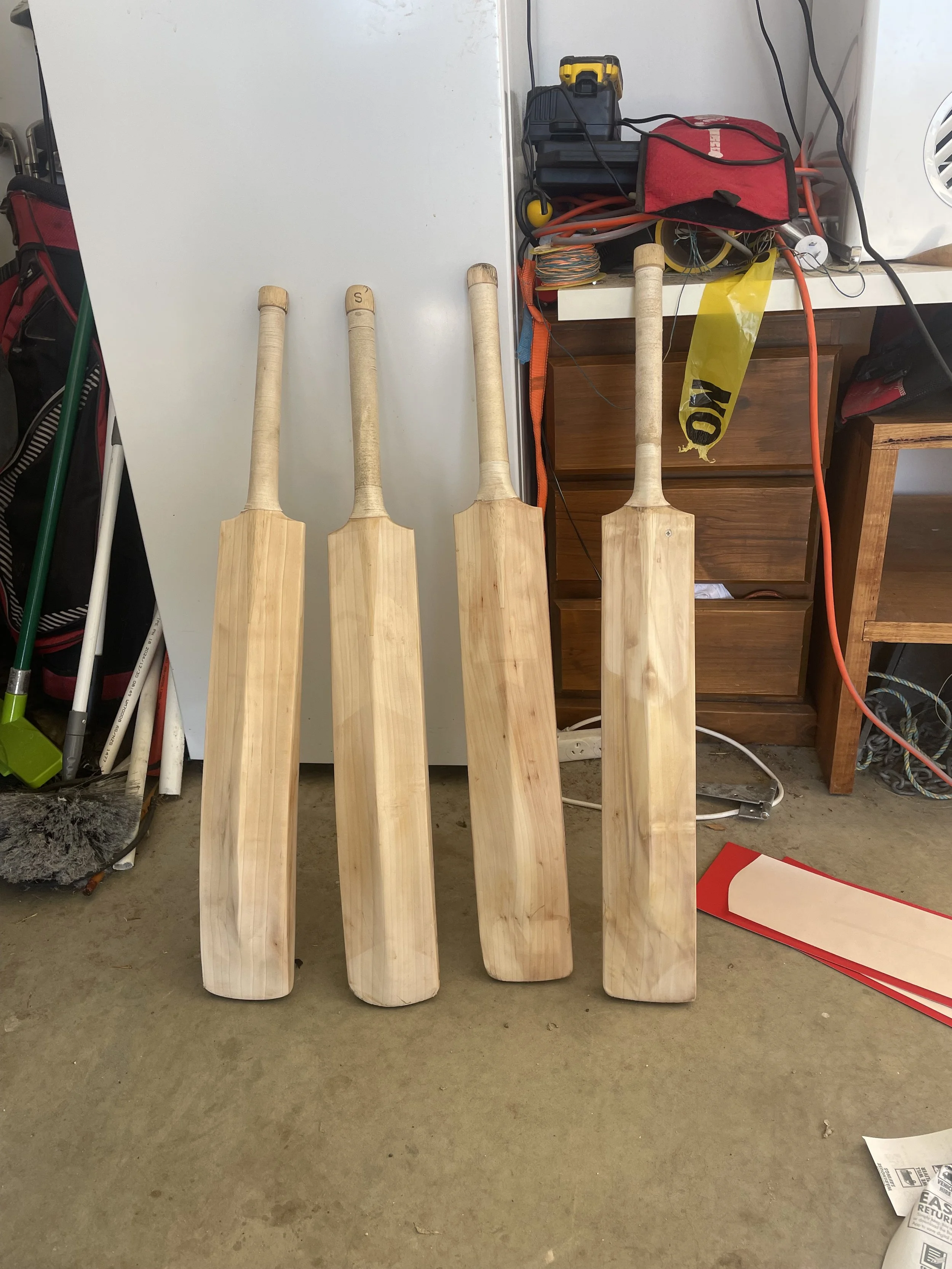Four wooden cricket bats leaning against a white surface in a garage or workshop.