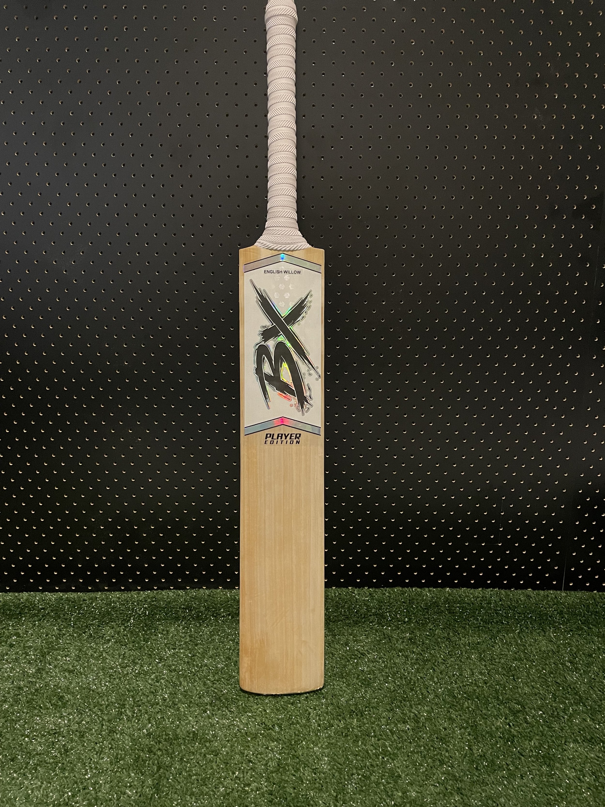 Cricket bat with a grip, displayed against a black perforated background and green artificial grass at the bottom.